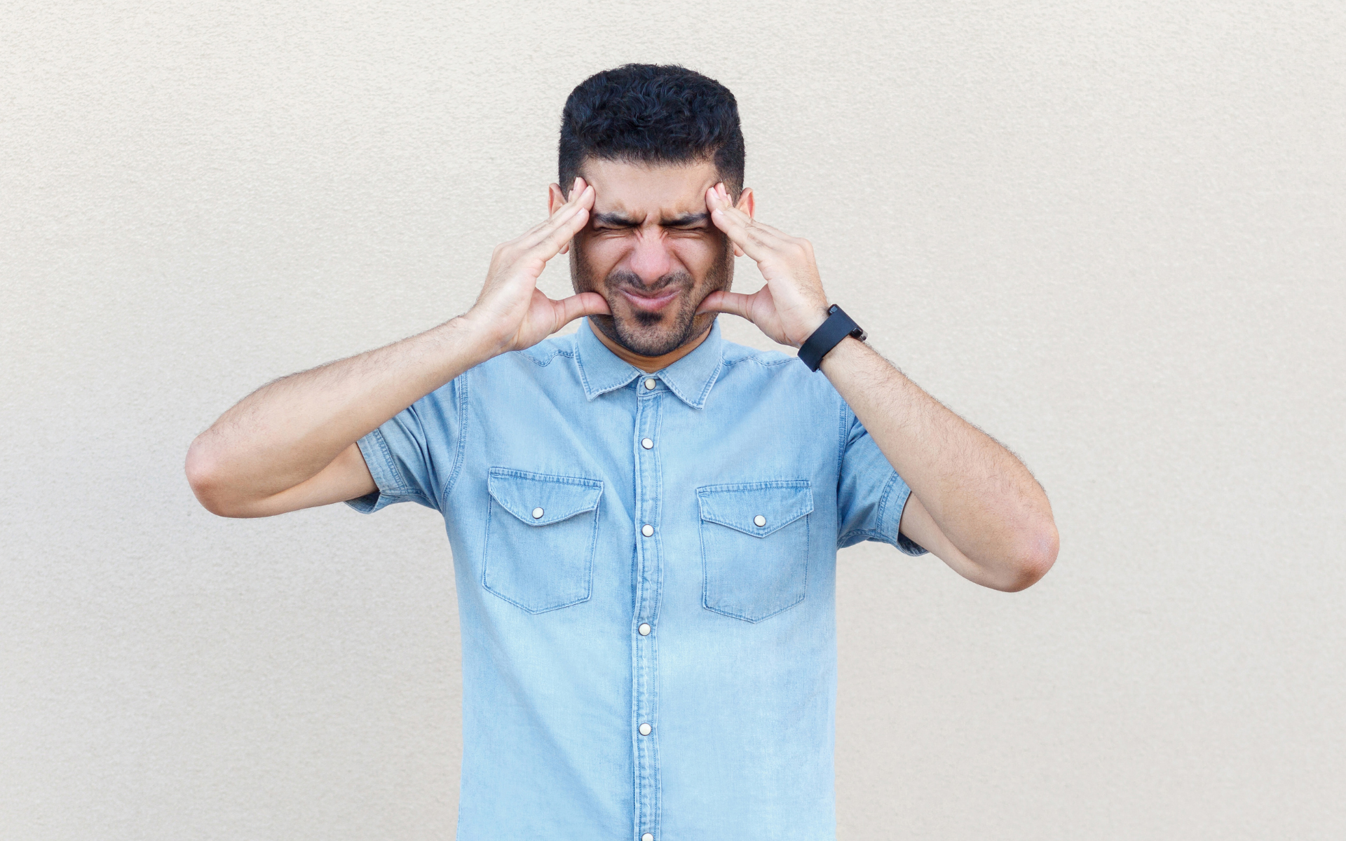 Man in a light blue shirt pressing his temples, looking stressed against a plain background