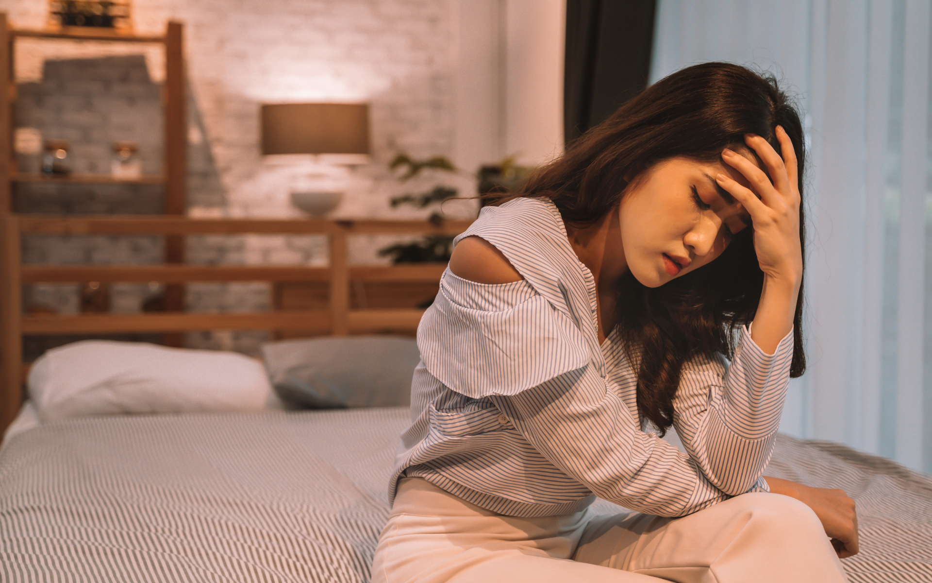 Woman sitting on a bed, holding her head in a bedroom, looking tired or upset
