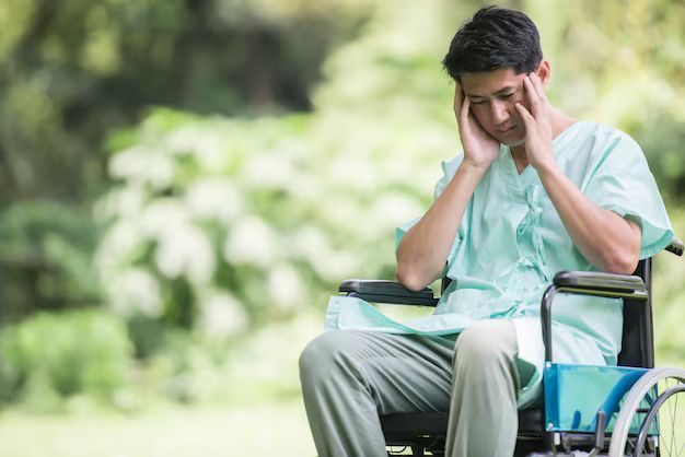 Man in a wheelchair holding his head, looking stressed outdoors with a blurred green background