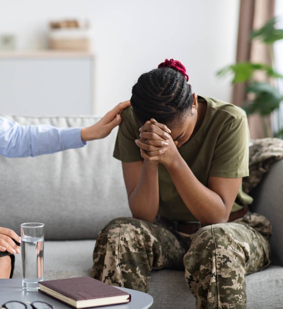 Person sitting on a couch, head in hands, while another offers comfort in a counseling setting