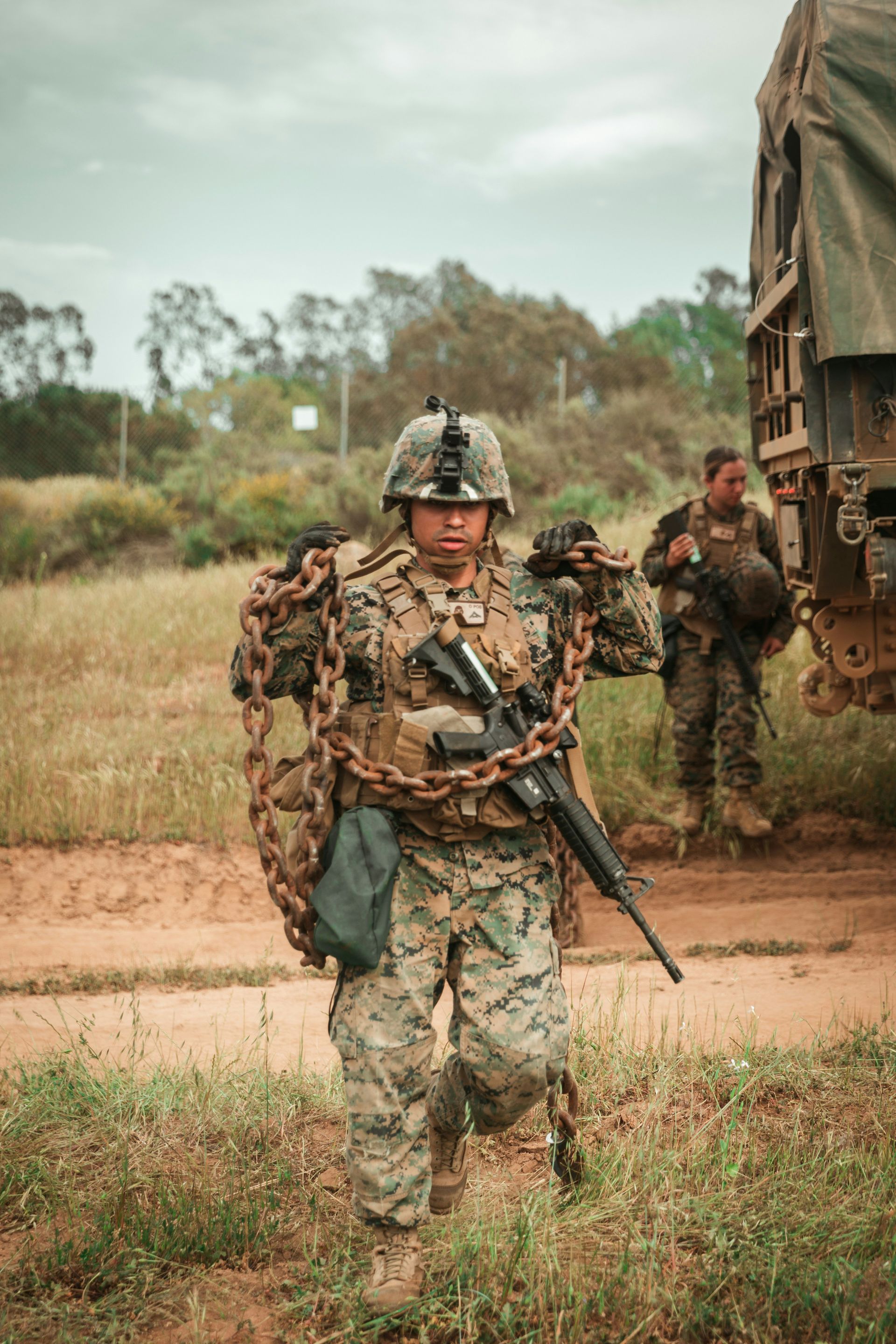 Camouflaged soldier holding a rifle, standing on a dirt path with military vehicles and other soldiers behind.