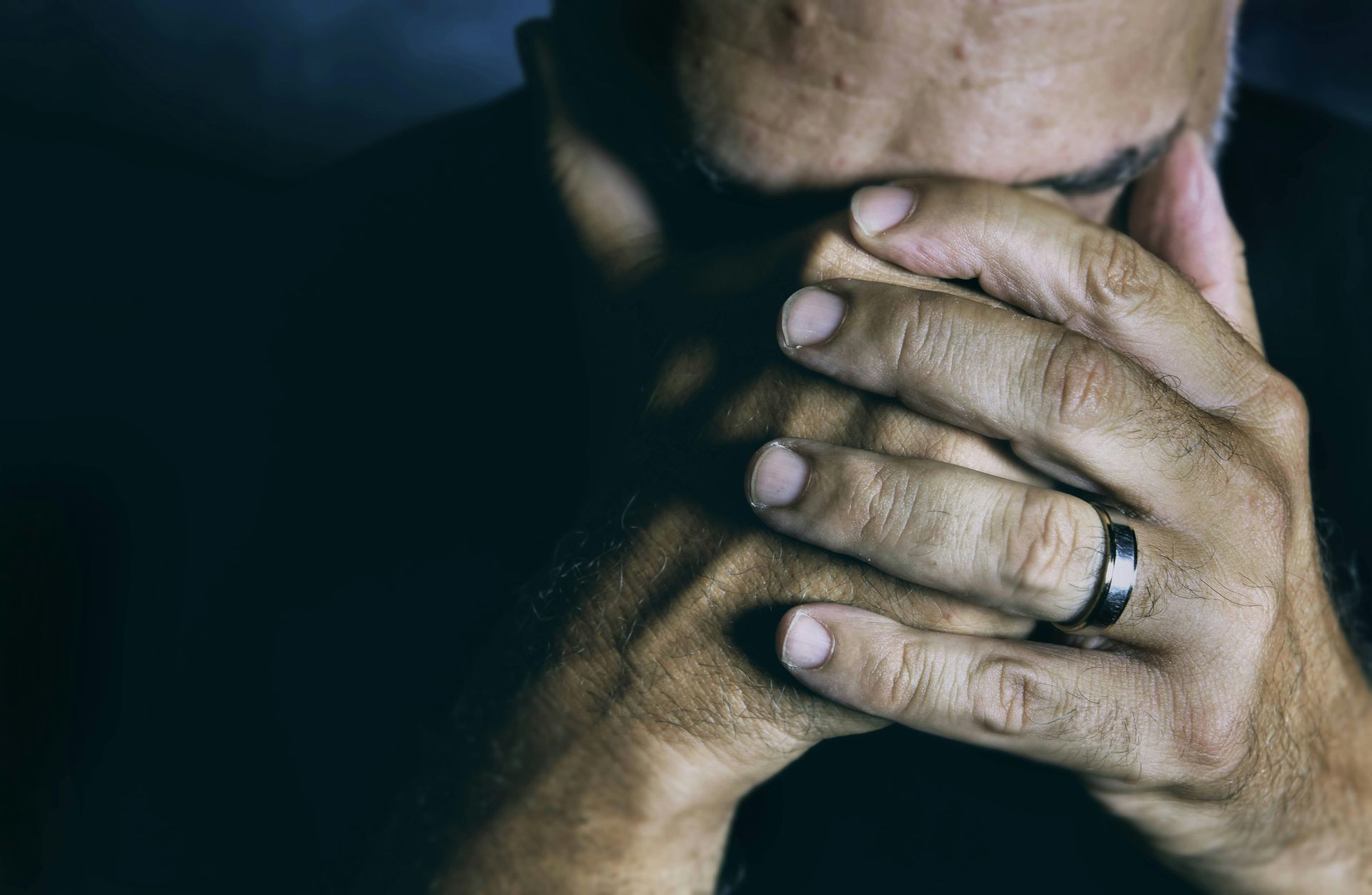 Close-up of a person covering their face with both hands, wearing a ring, against a dark background