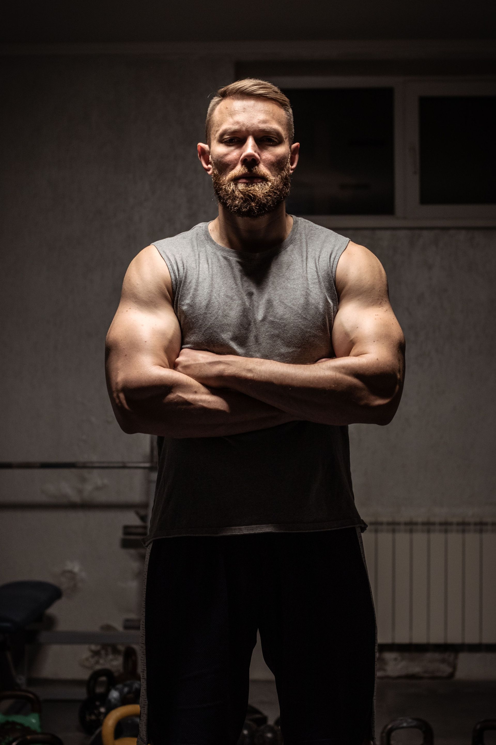 Muscular man with arms crossed in a dim gym, wearing a gray sleeveless shirt.