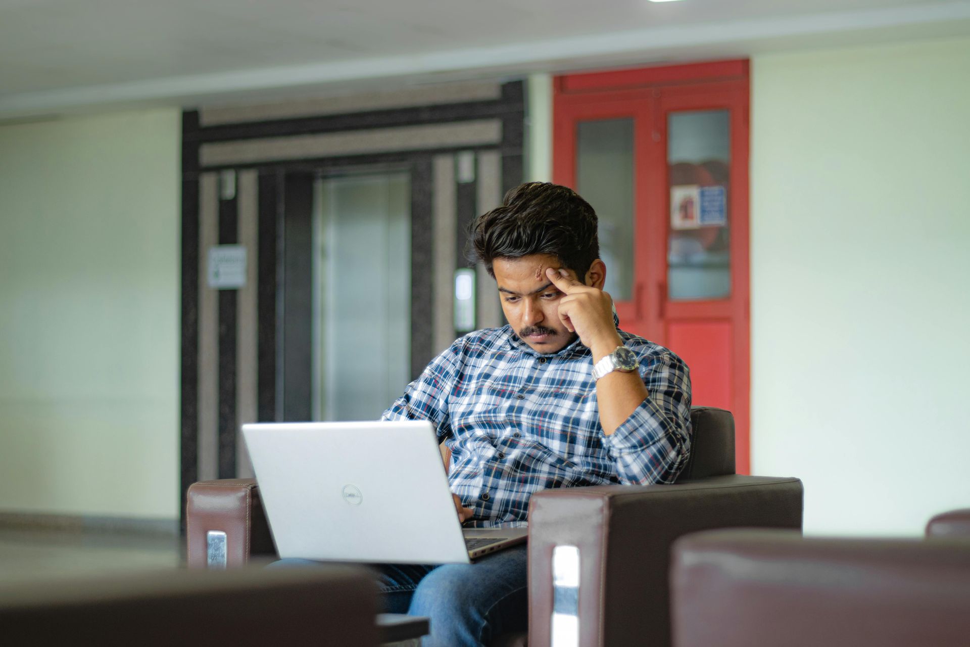 Man seated in a lounge, working on a laptop with a hand to his forehead, looking stressed.