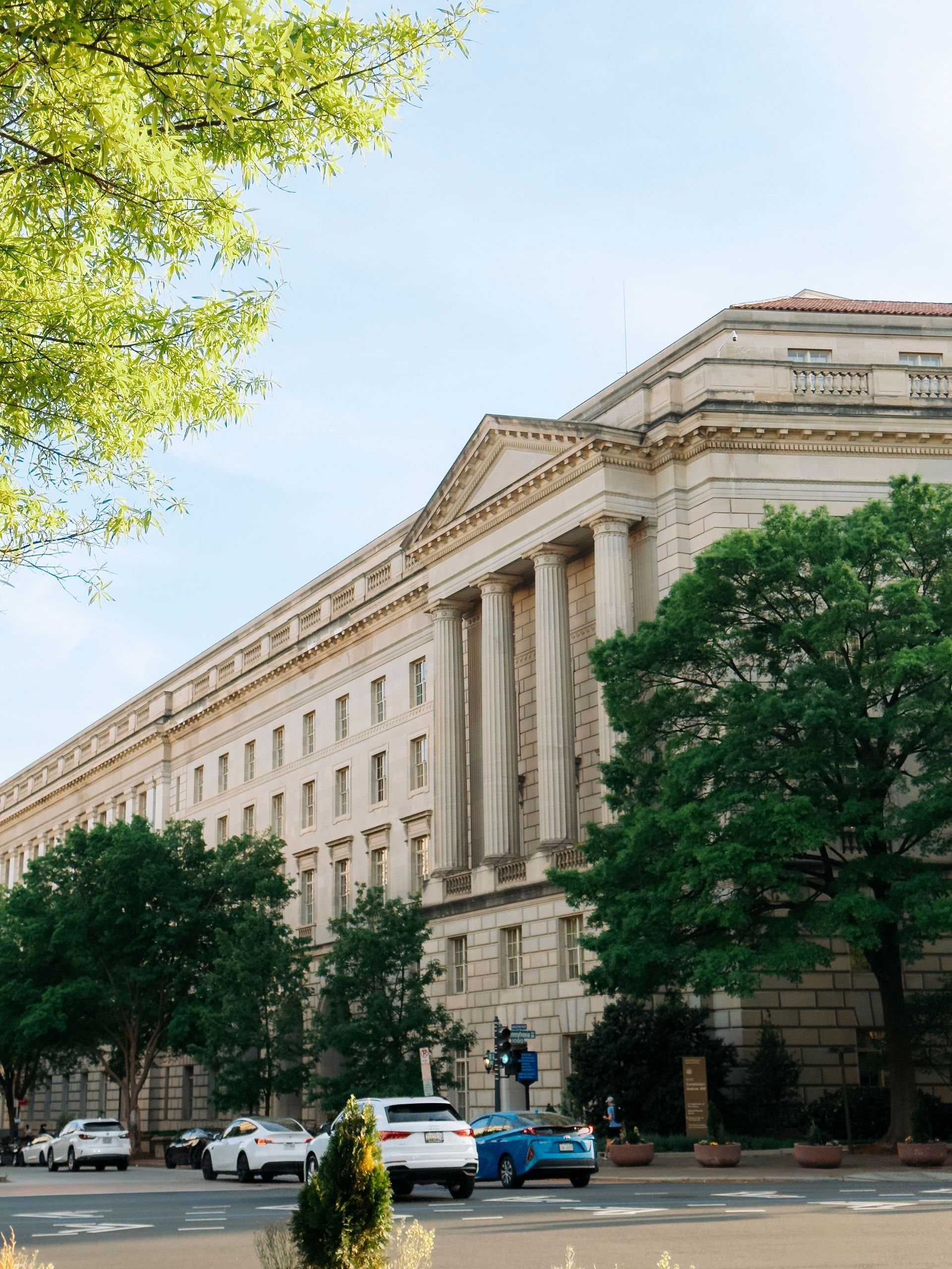 Classical stone government building with columns, trees, and parked cars on a sunny street