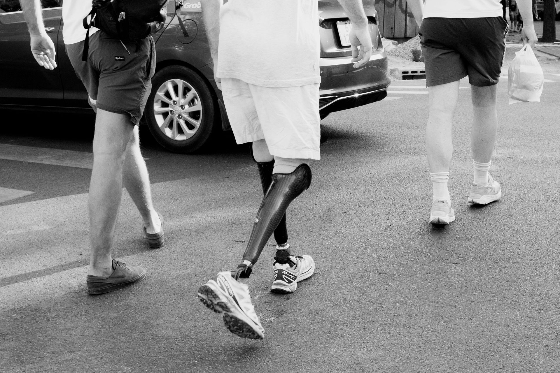 Three people walking on a street past parked cars, shown in black and white.