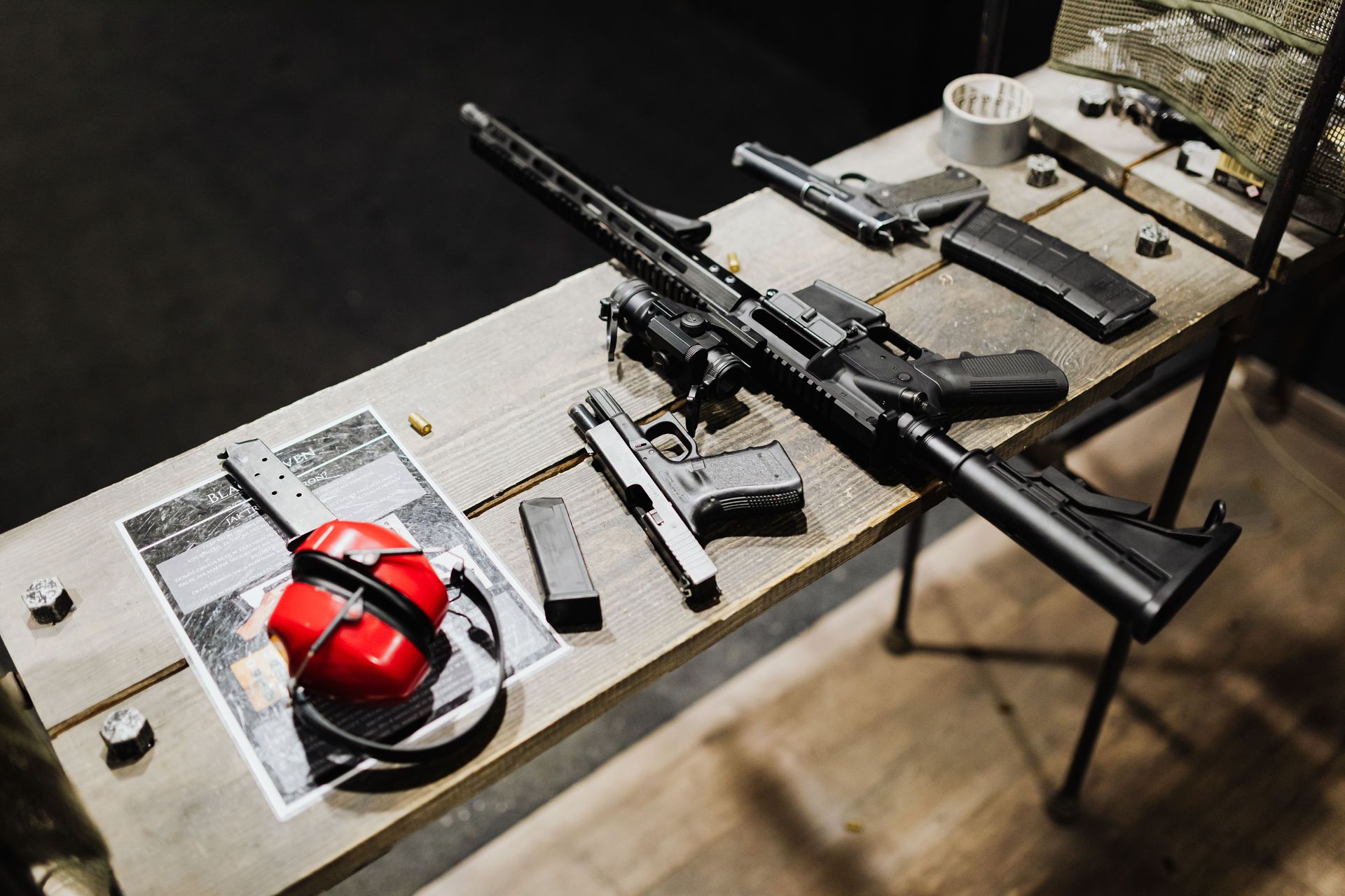 Rifle and handgun displayed on a table beside red hearing protection in an indoor shooting range