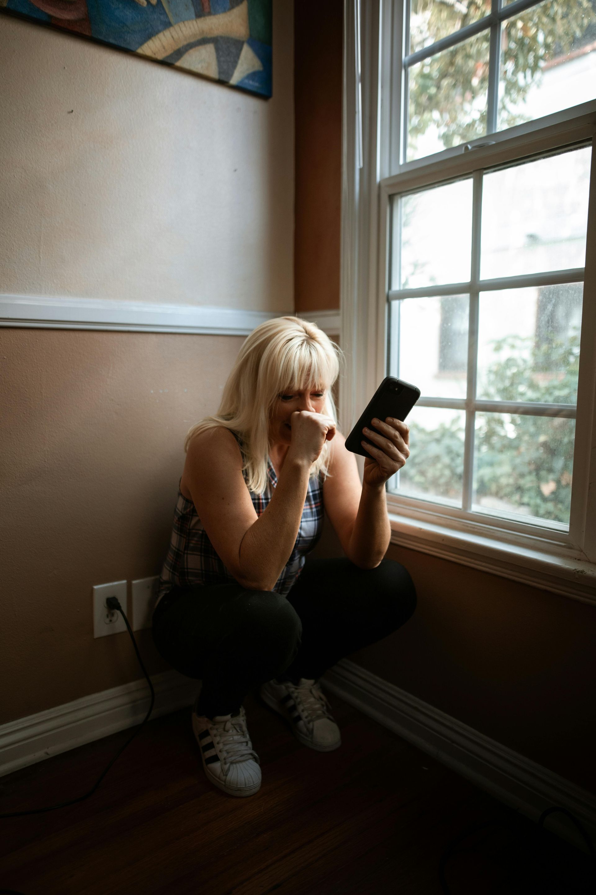 Person crouches by a sunlit window, looking at a phone in a small room.