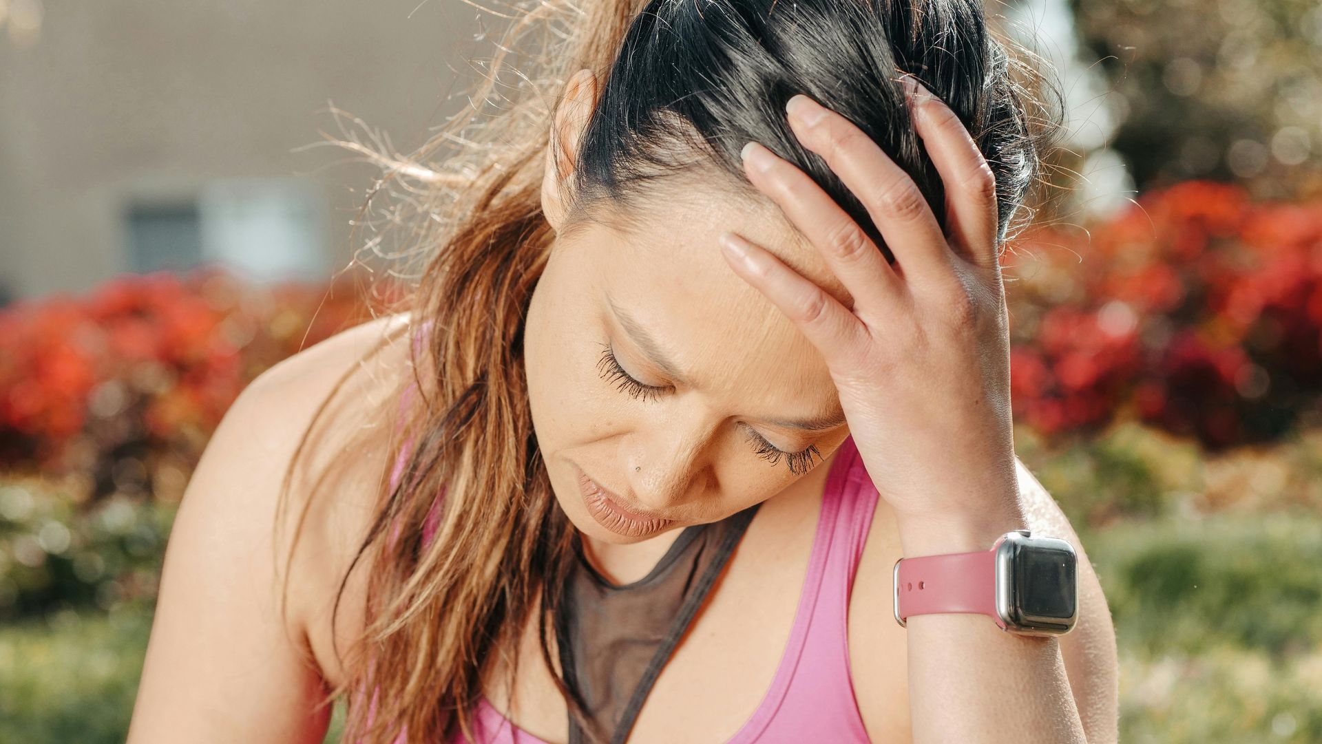 Woman outdoors, holding her head with a pink fitness watch on her wrist, looking fatigued.