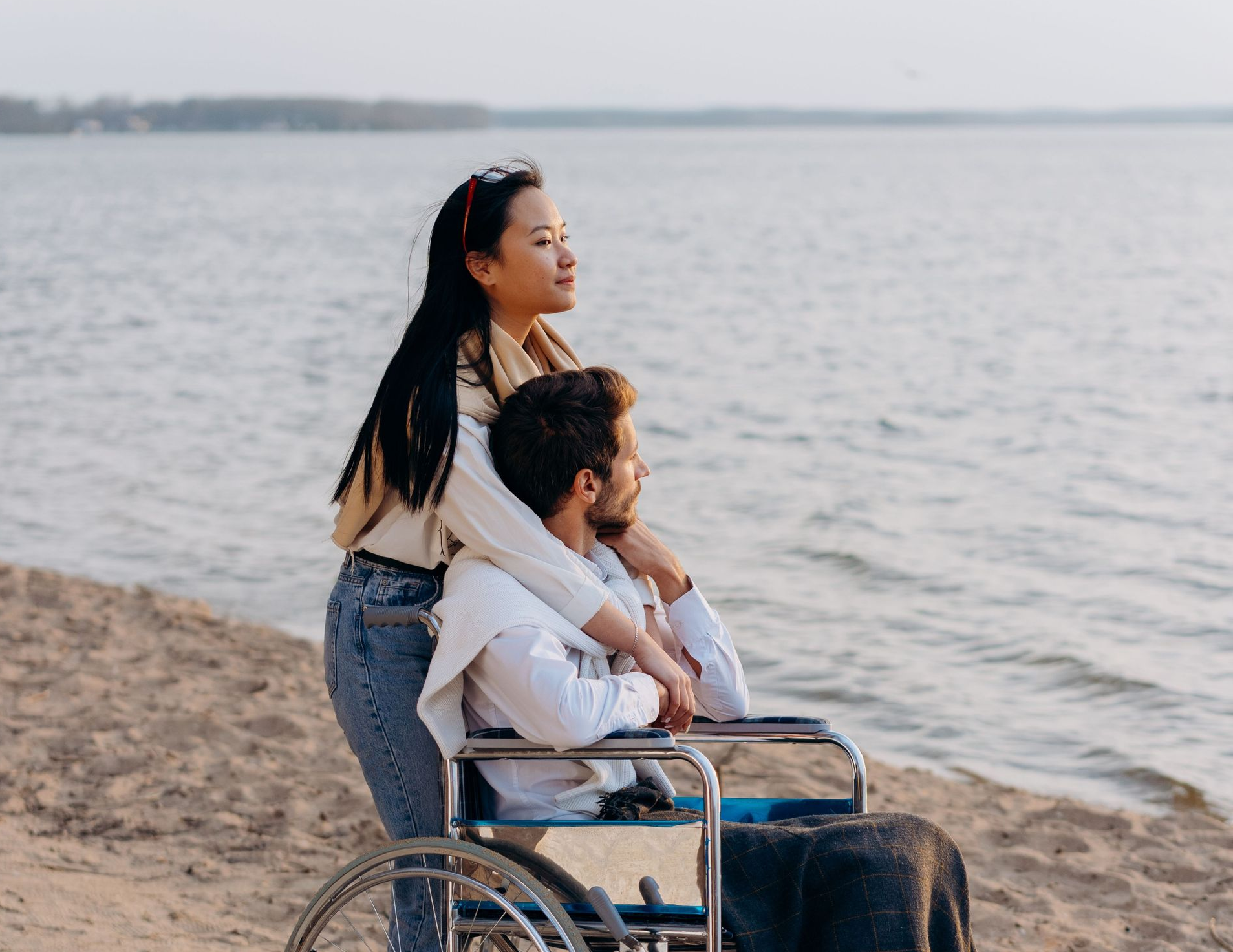 Two people by the water; one stands behind a person in a wheelchair on the sandy shore.