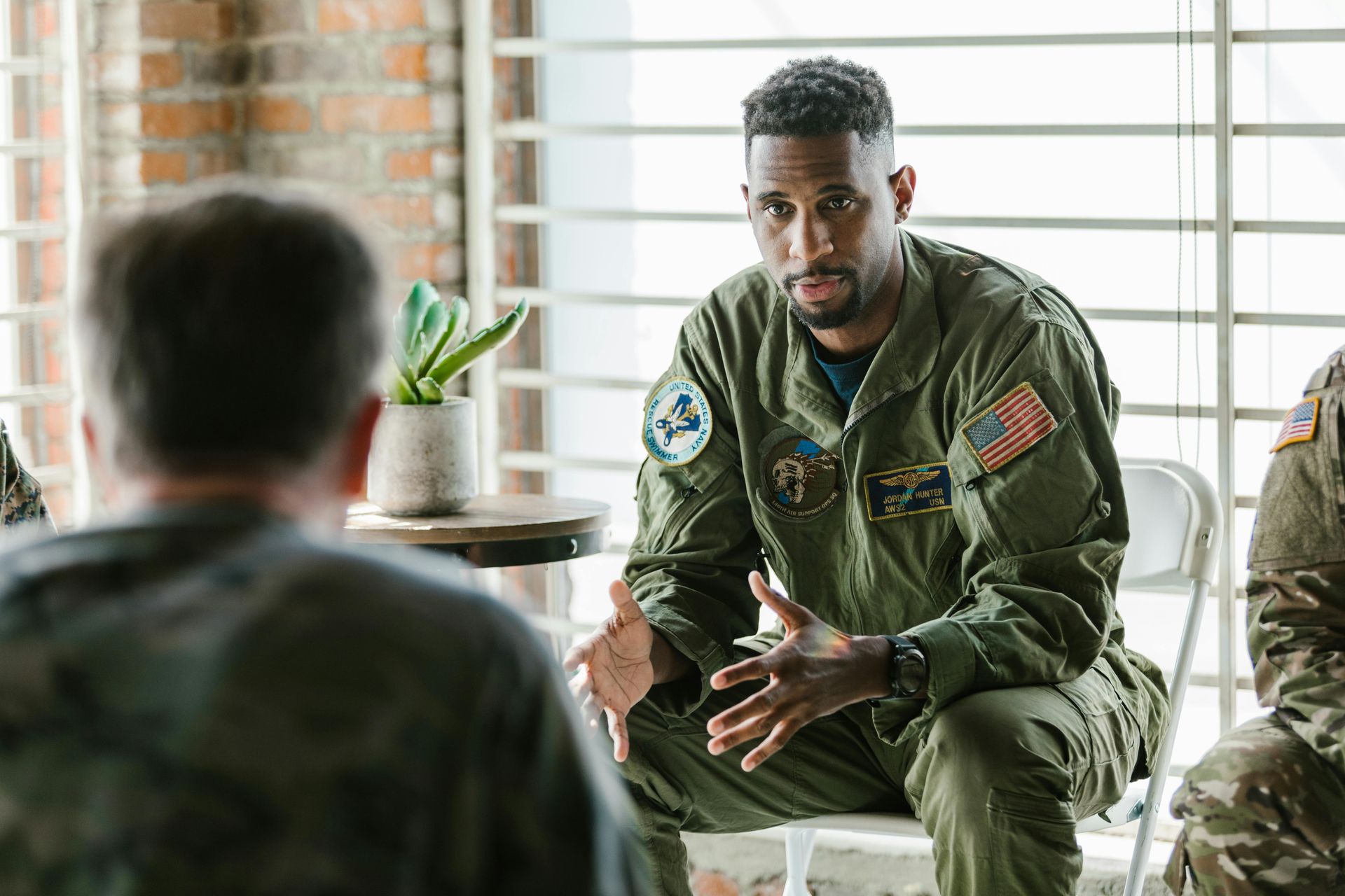 Military officer in green flight suit speaking indoors, seated by a window with blinds.