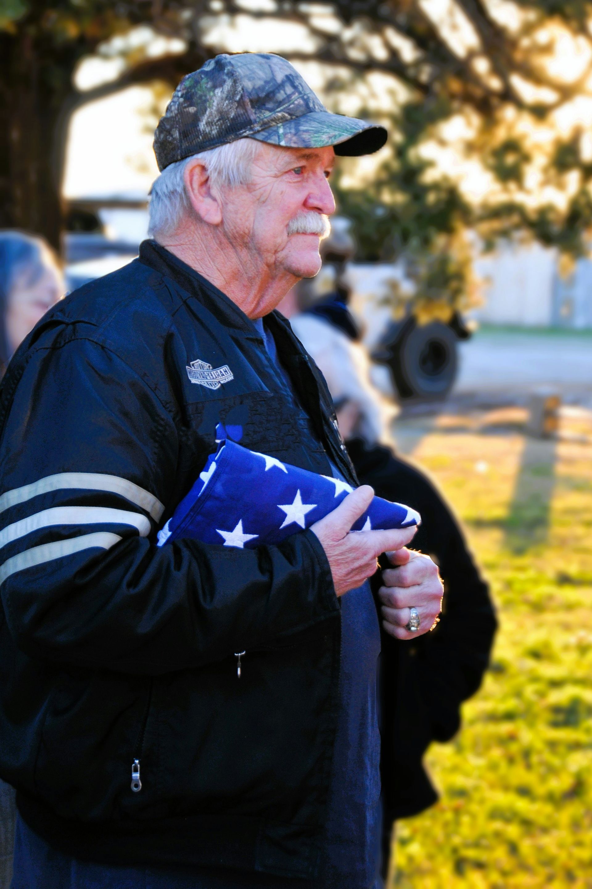 Man holding a folded American flag outdoors at sunset, wearing a dark jacket and camouflage cap
