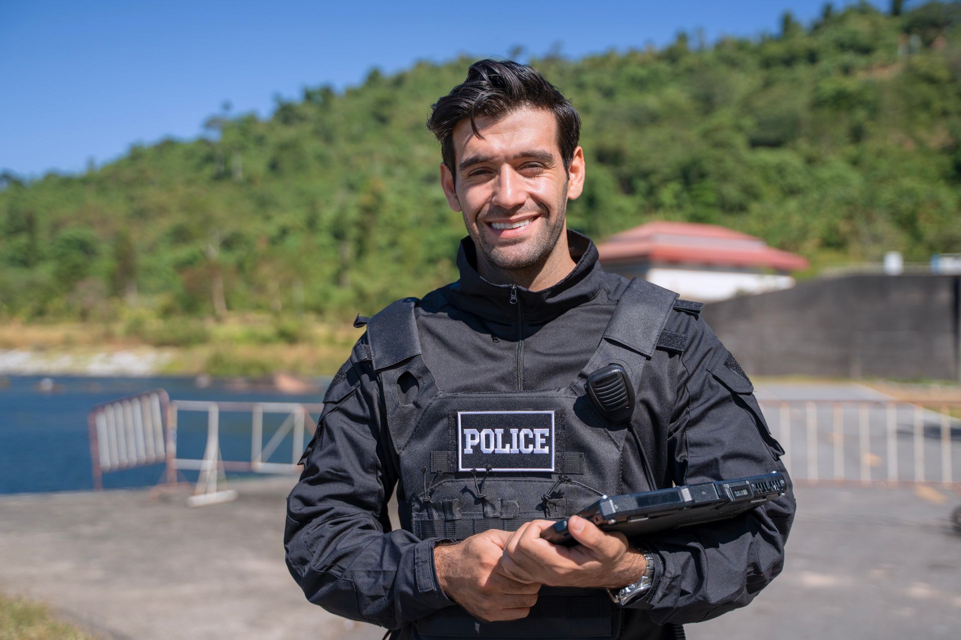 Police officer in black tactical gear holding a rifle outdoors near a road barrier and hillside