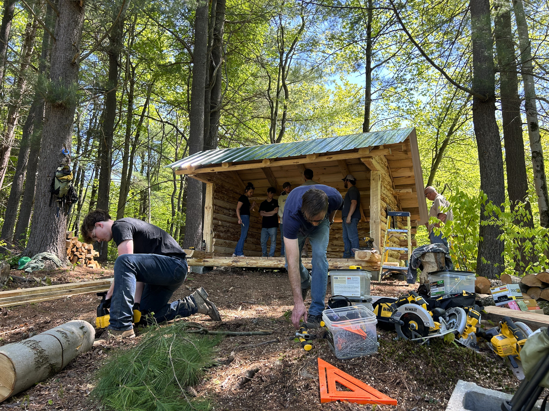 People building a lean-to in a wooded forest clearing with tools and construction materials nearby