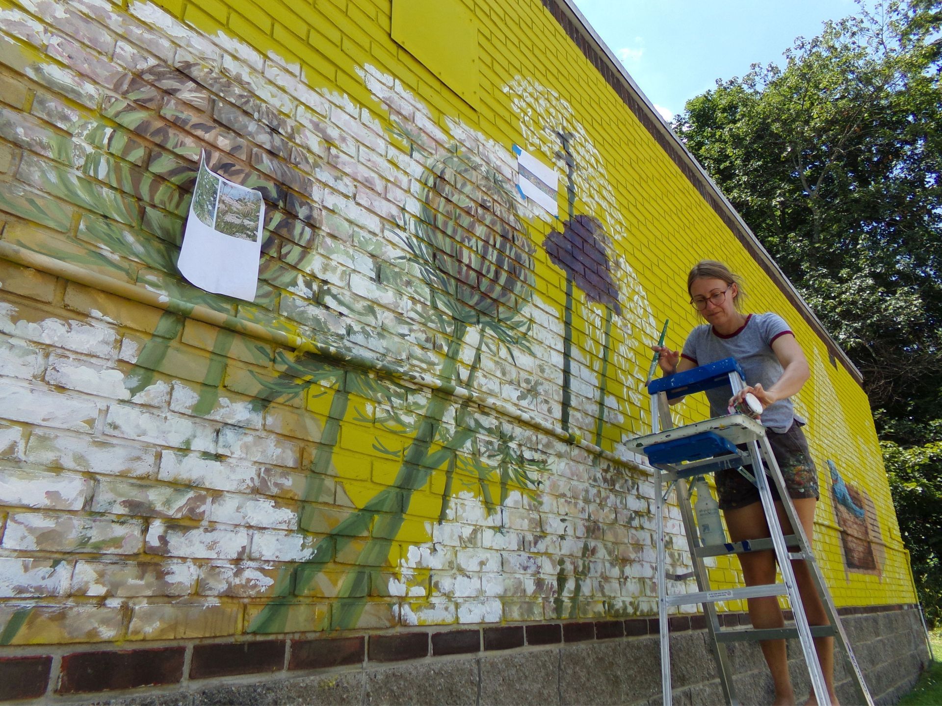 Person painting a colorful mural on a brick wall while standing on a ladder
