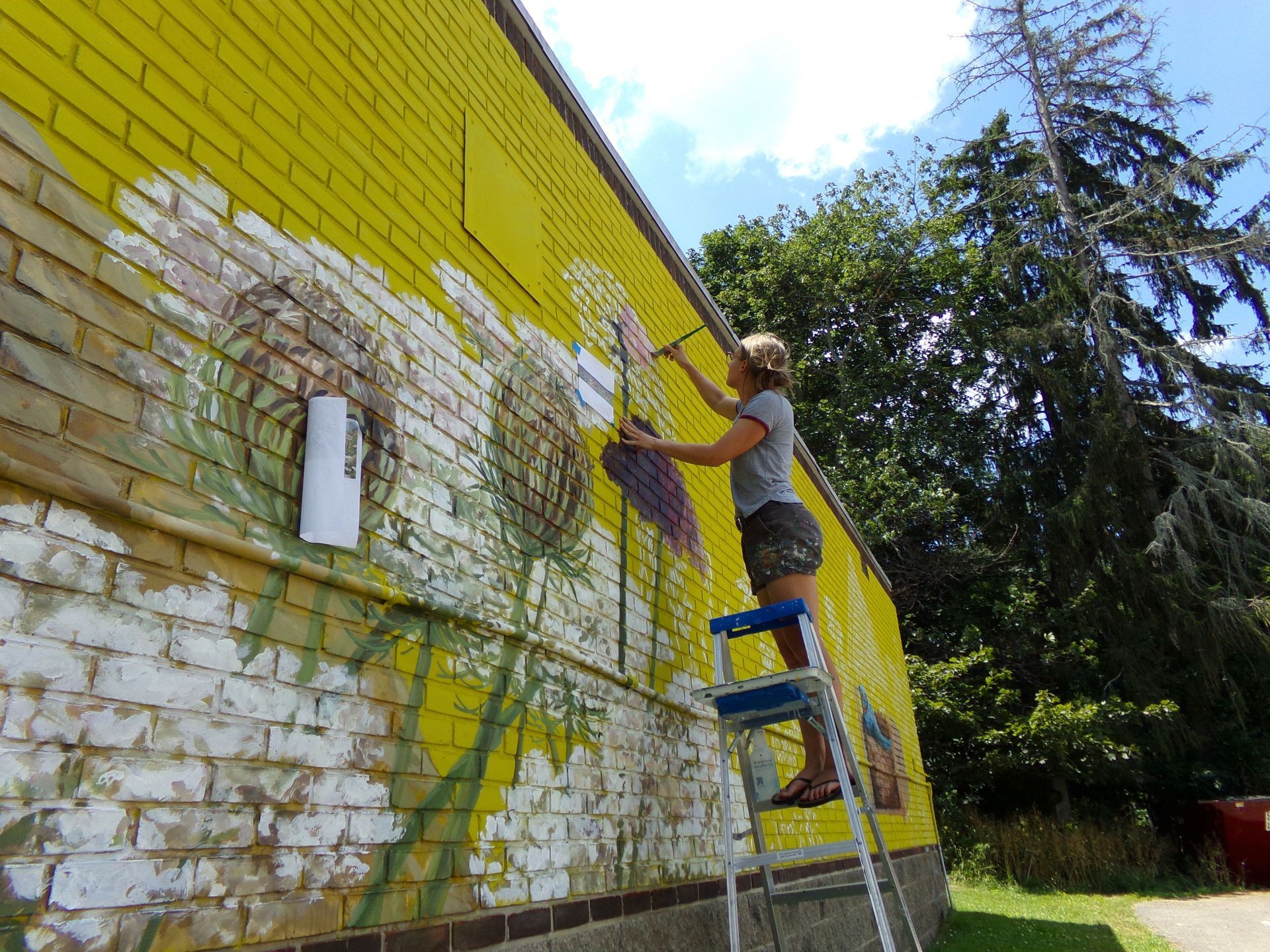 Person on ladder painting a brick wall bright yellow outdoors