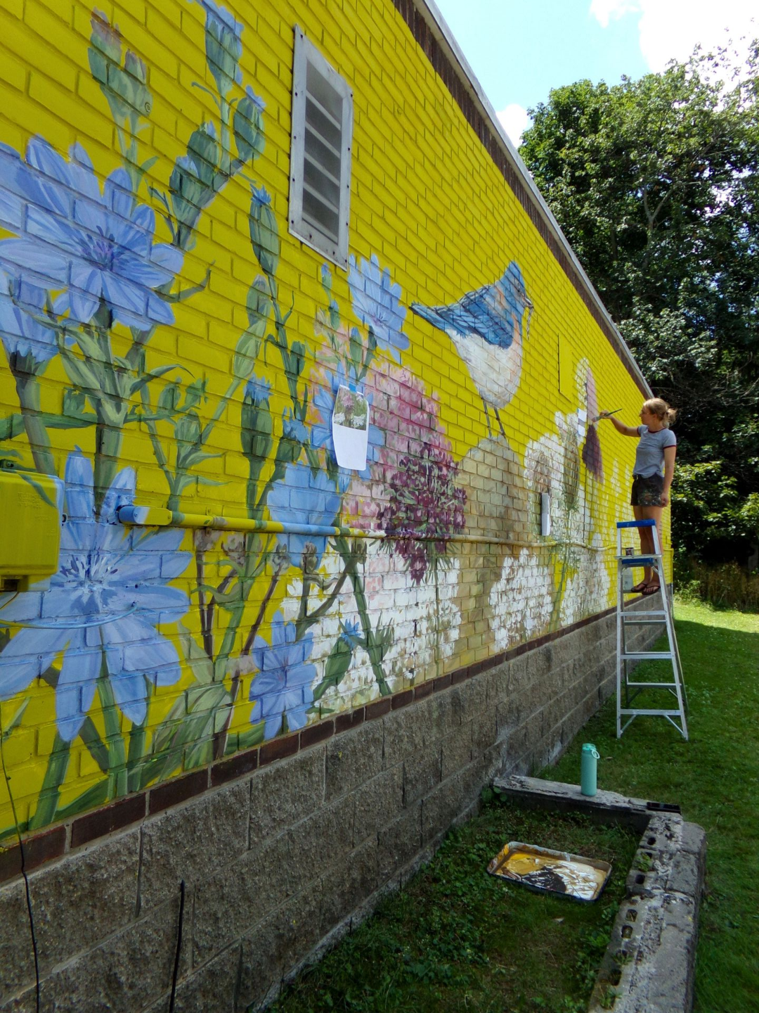 Chloe Smith painting a colorful mural on a yellow brick wall outdoors while standing on a ladder