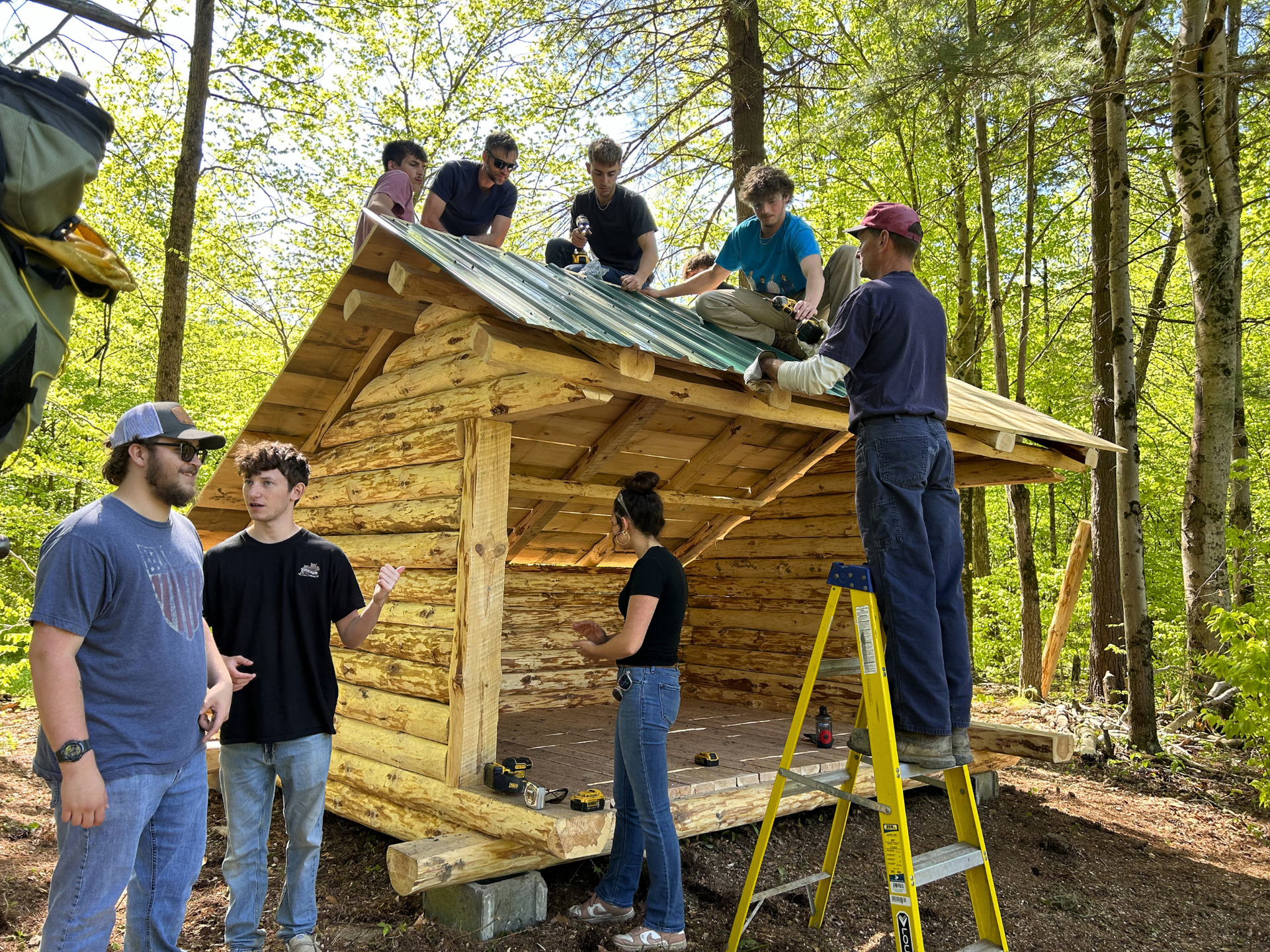 People building a wooden lean-to on West Almond Trail, with several standing on the roof and one on a ladder.