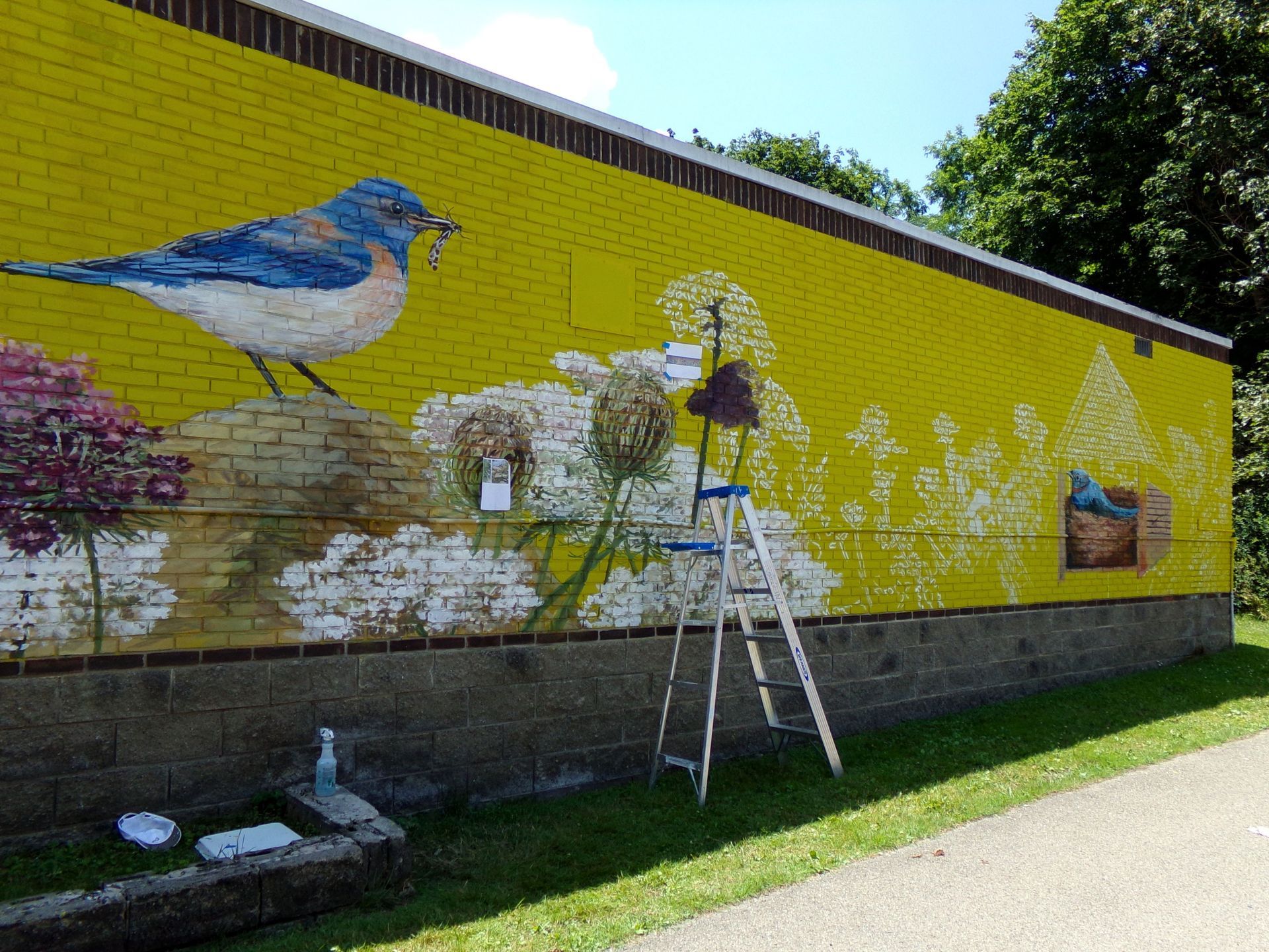 Person on ladder painting a yellow brick wall mural with birds and flowers outdoors.