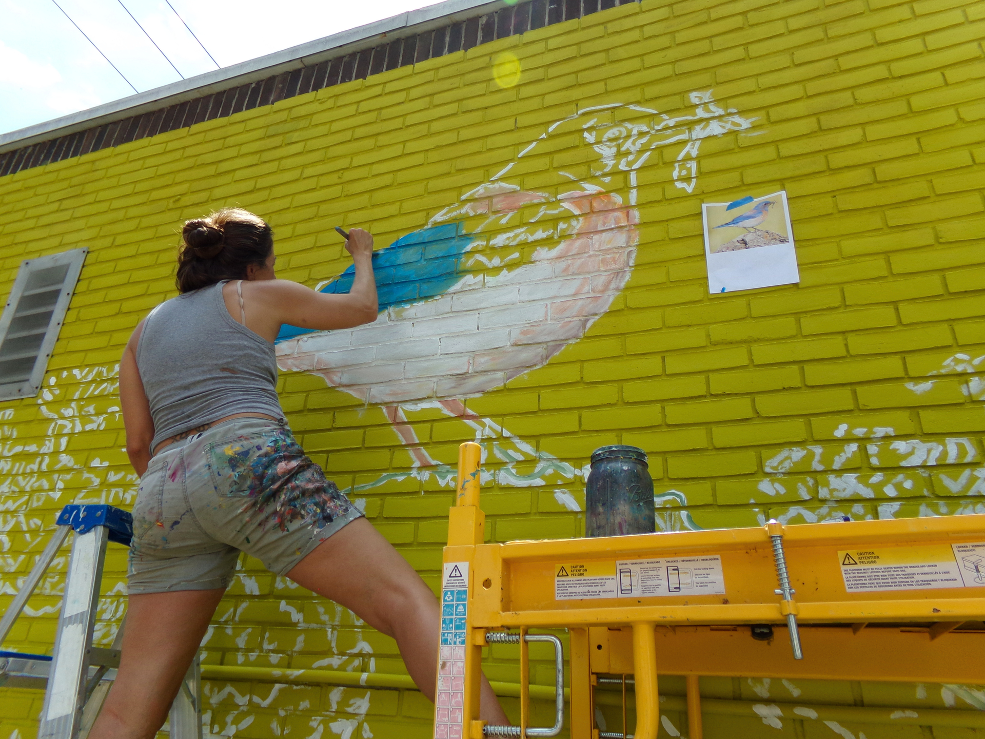 Person painting a mural on a yellow brick wall beside a yellow scaffolding table