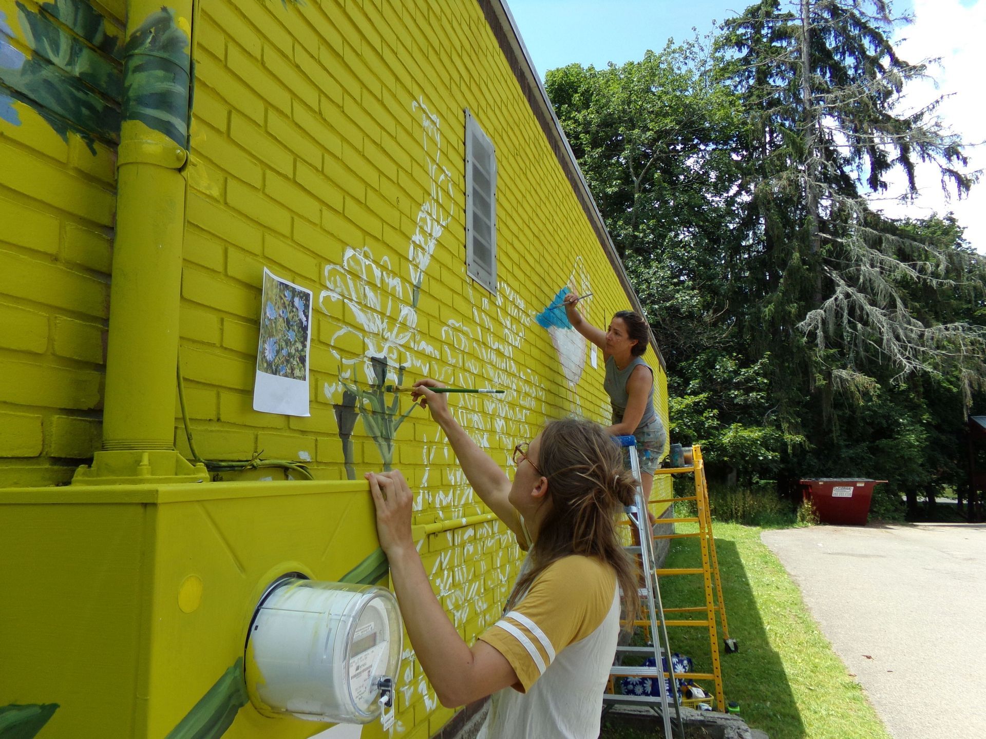 Two people painting a bright yellow brick wall outdoors with a ladder.