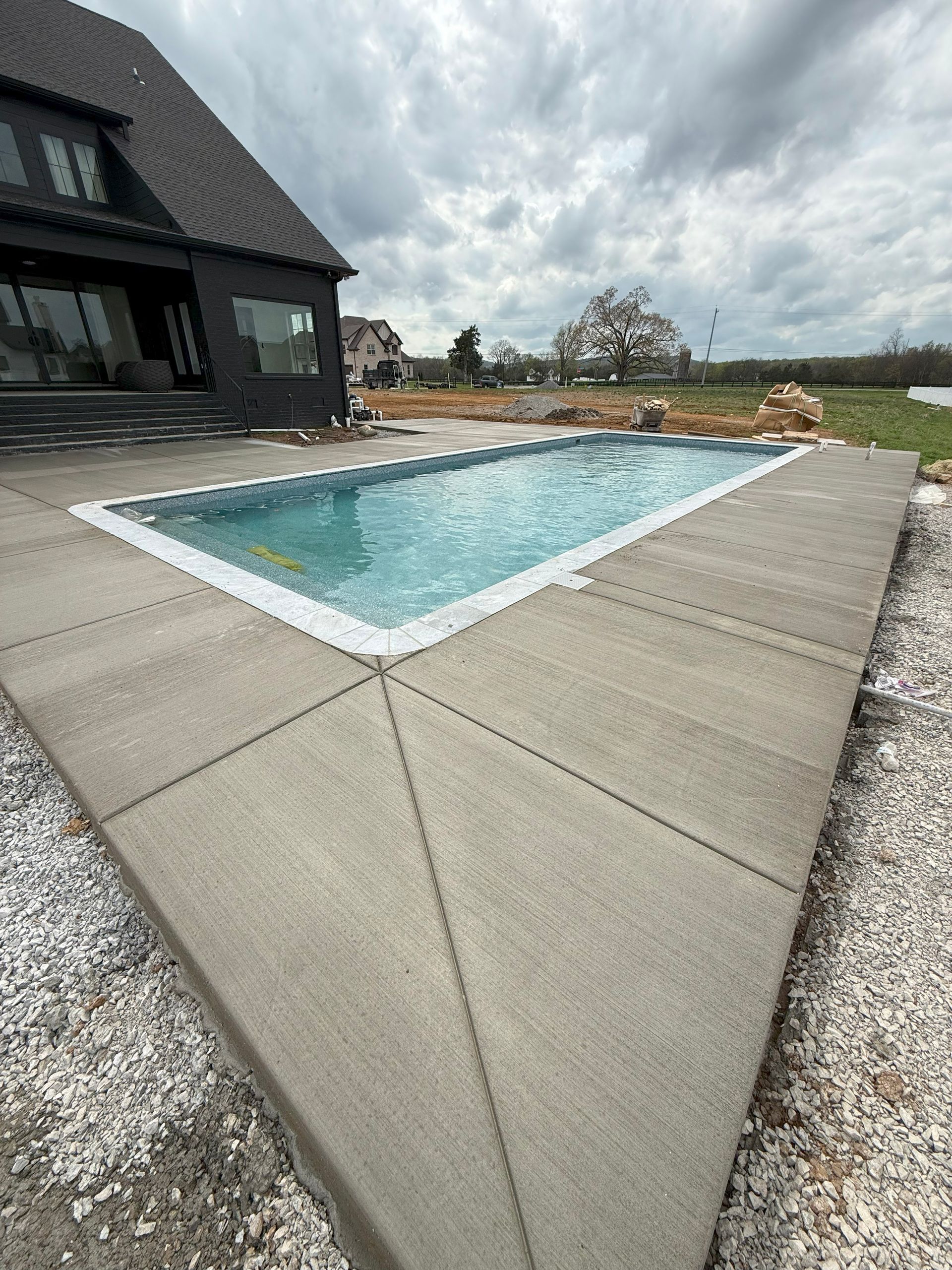 Rectangular backyard pool beside a dark house, with a wide concrete patio and cloudy sky.