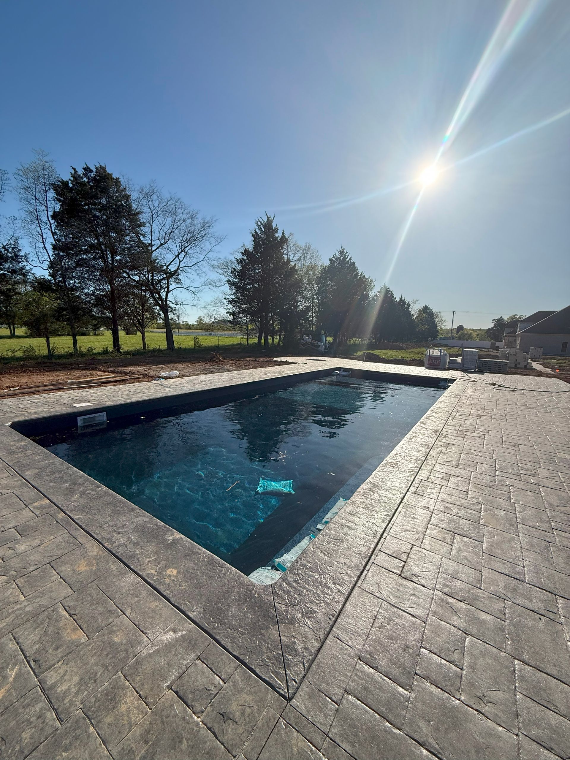 Stone patio with a small rectangular pool reflecting a bright sun and trees under a clear blue sky