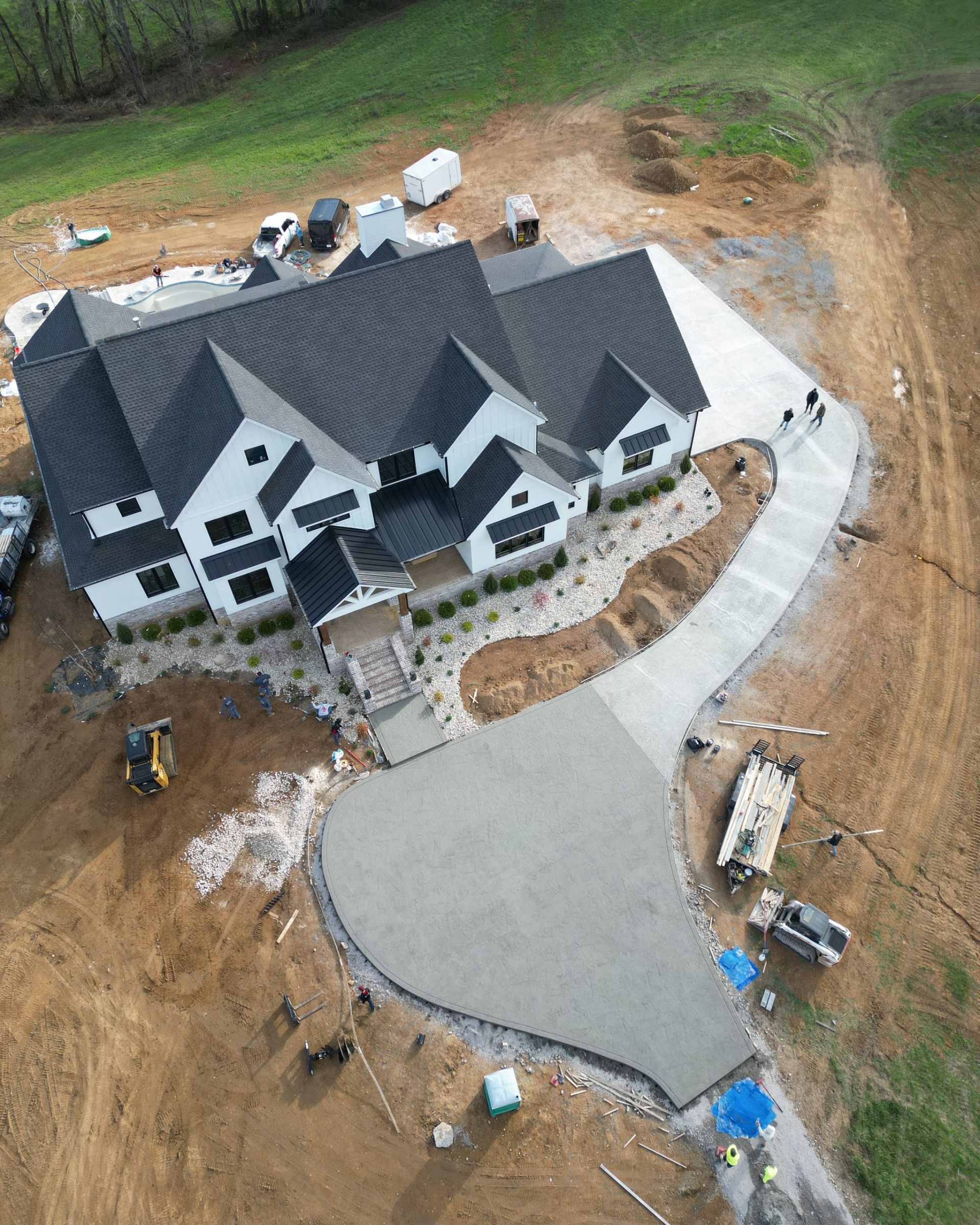 Aerial view of a large house under construction with a curved concrete driveway and surrounding dirt lot.