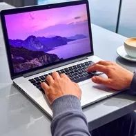 View Web Features (pop up) Hands typing on a laptop with a purple mountain wallpaper on a desk beside a cup of coffee.