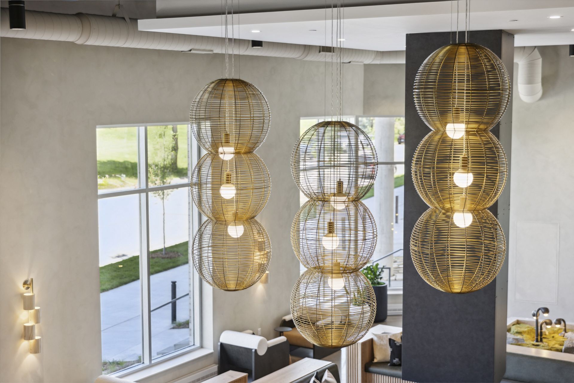 Three light fixtures with stacked, open-sphere cages hang from the ceiling. A window and interior architecture are in the background.