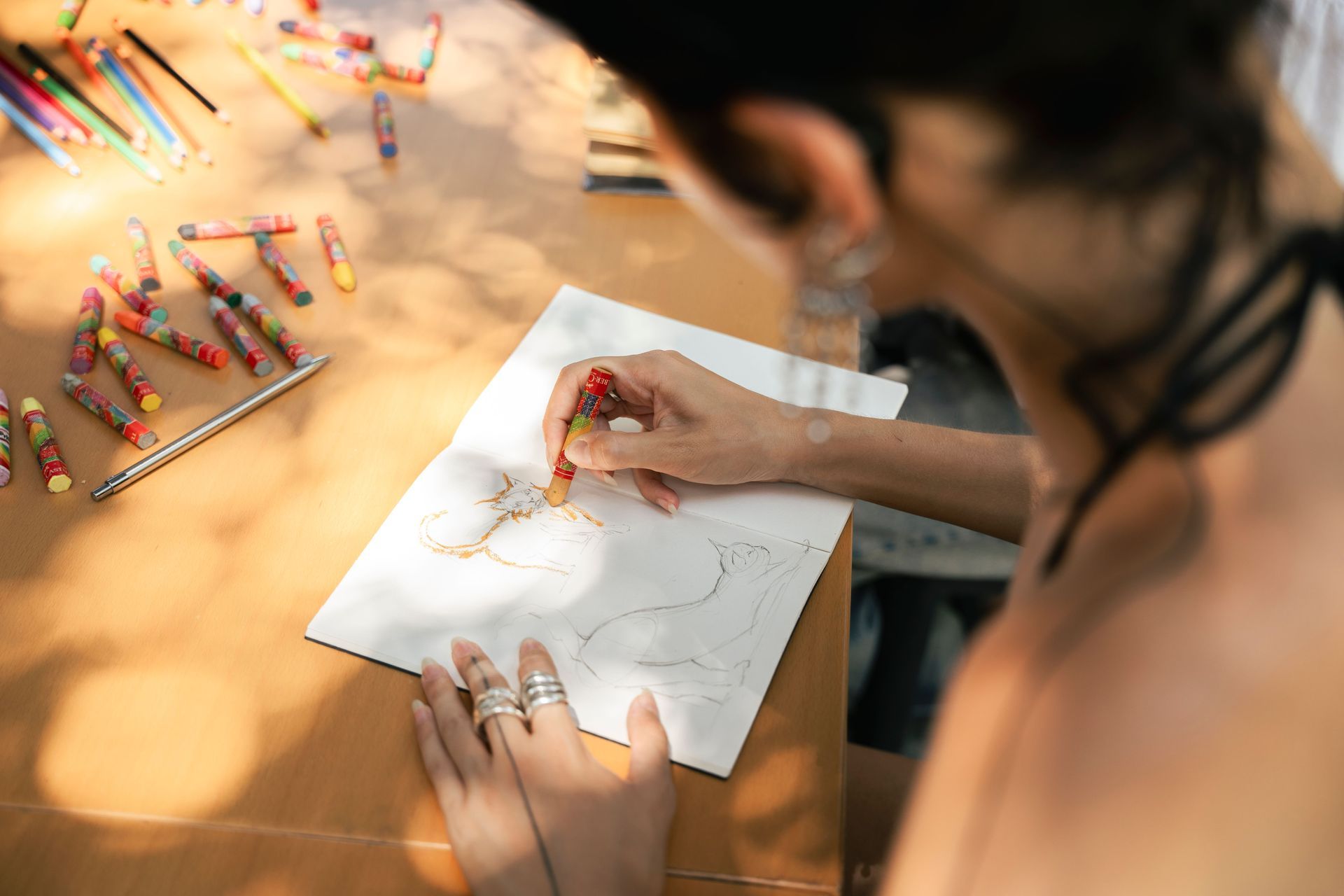 Person drawing with a red pencil on white paper, with colorful pencils scattered on a table.