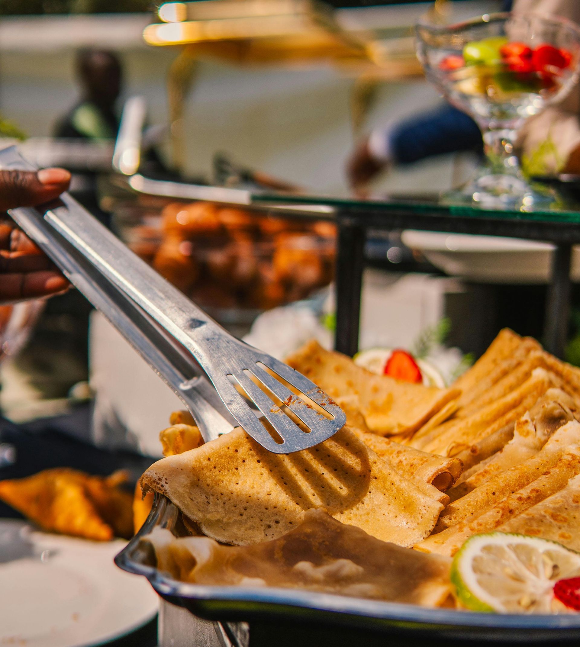 Tongs serving grilled fish fillets on a buffet tray with lemon slices and salad in the background