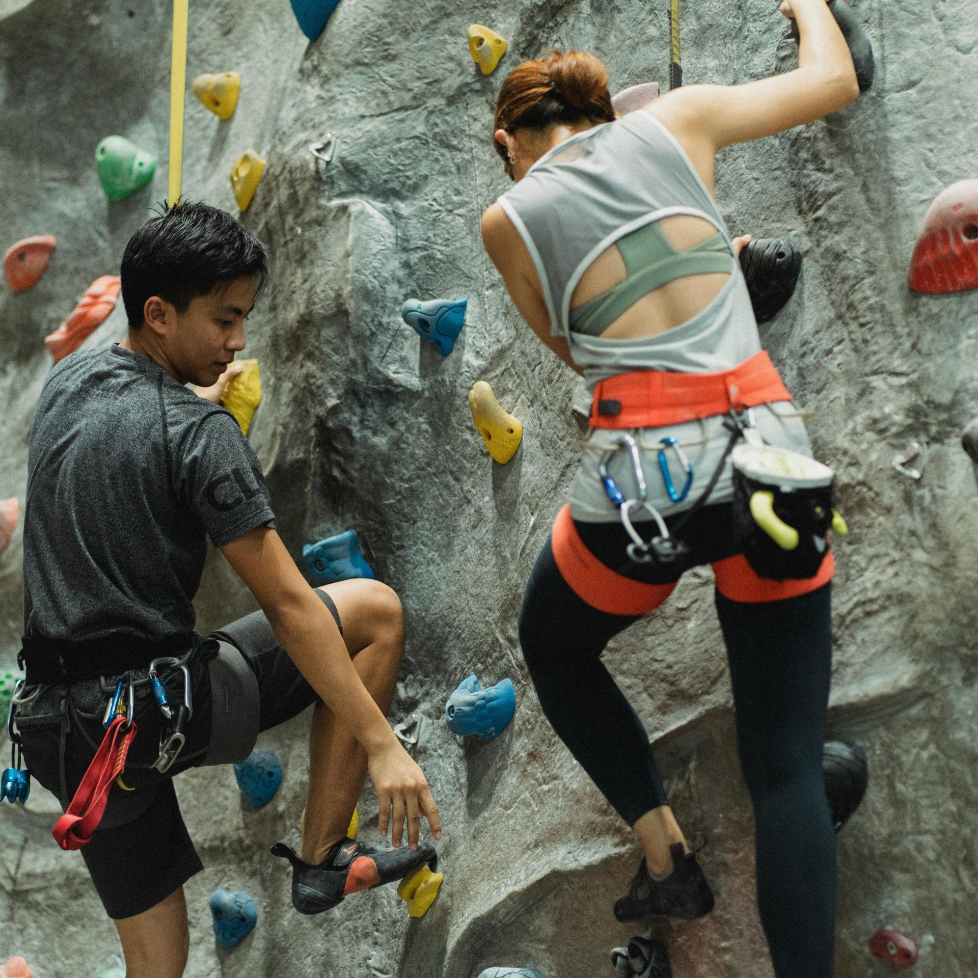 Two people rock climbing on an indoor wall. Both wear harnesses and athletic clothing.