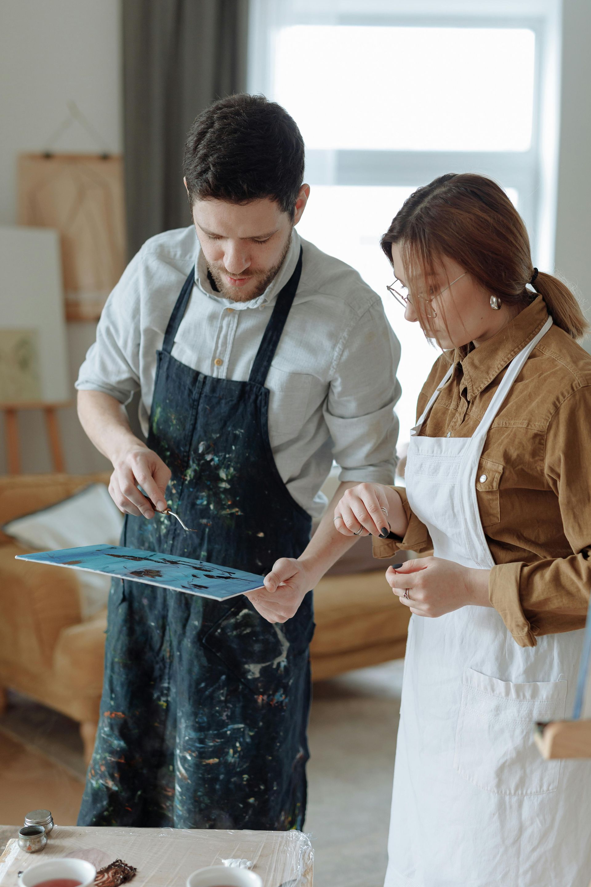 Two people wearing aprons study a paint-splattered palette while working together in a sunlit art studio.