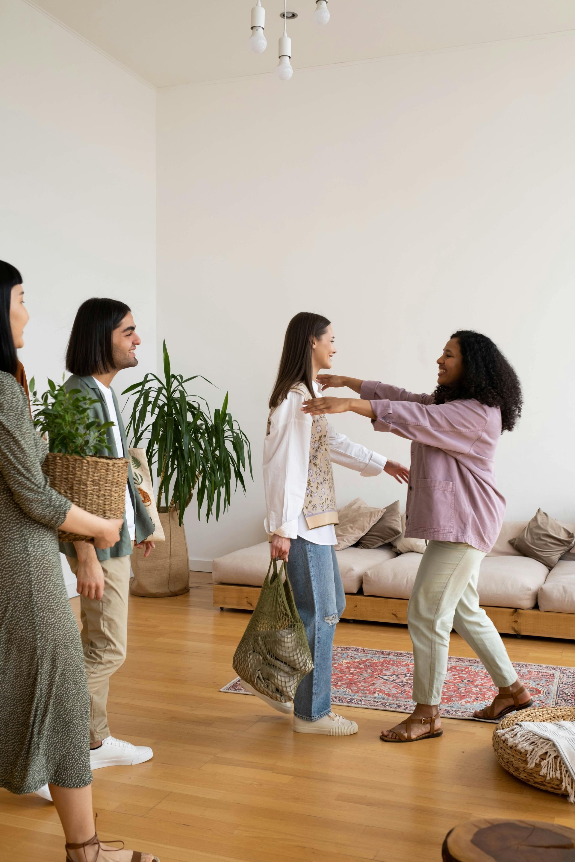 Two people greet each other with a hug in a bright living room, while two others stand nearby holding house plants.