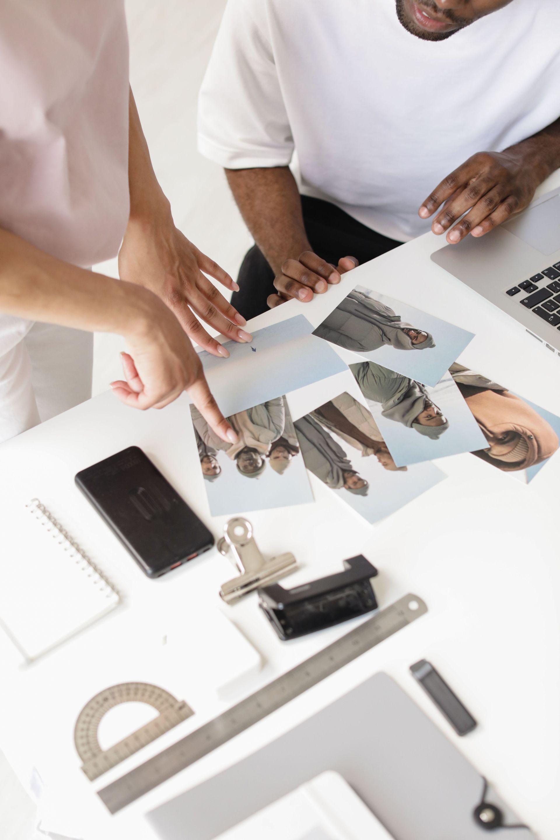 Hands reviewing design sketches on a white desk with tools, smartphone, and laptop keyboard