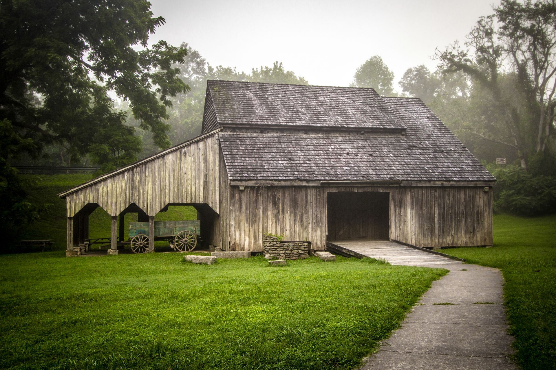 Old wooden barn with a thatched roof beside a curved path in a misty green field