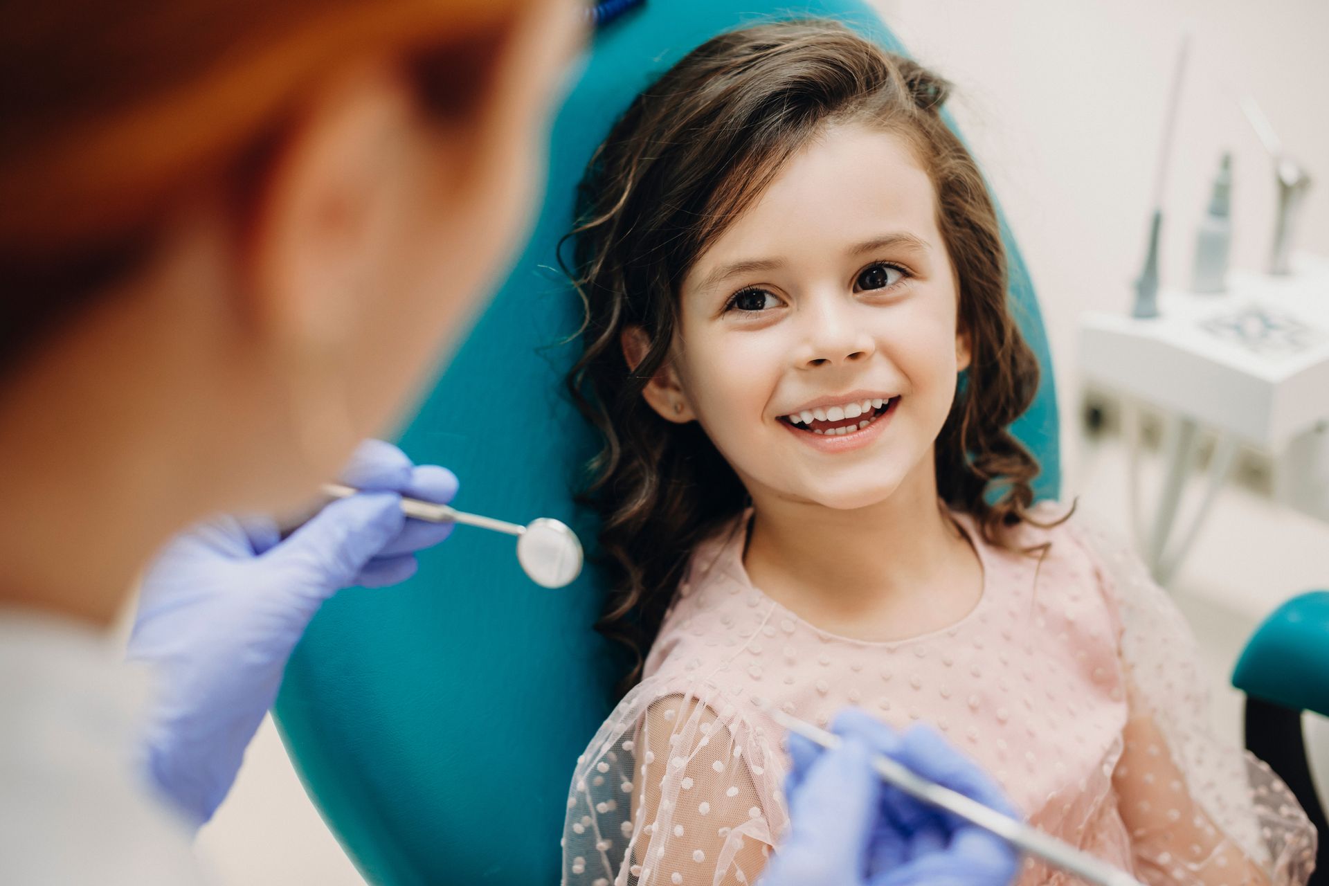 A smiling child sits in a dental chair while a dentist holds a dental mirror.