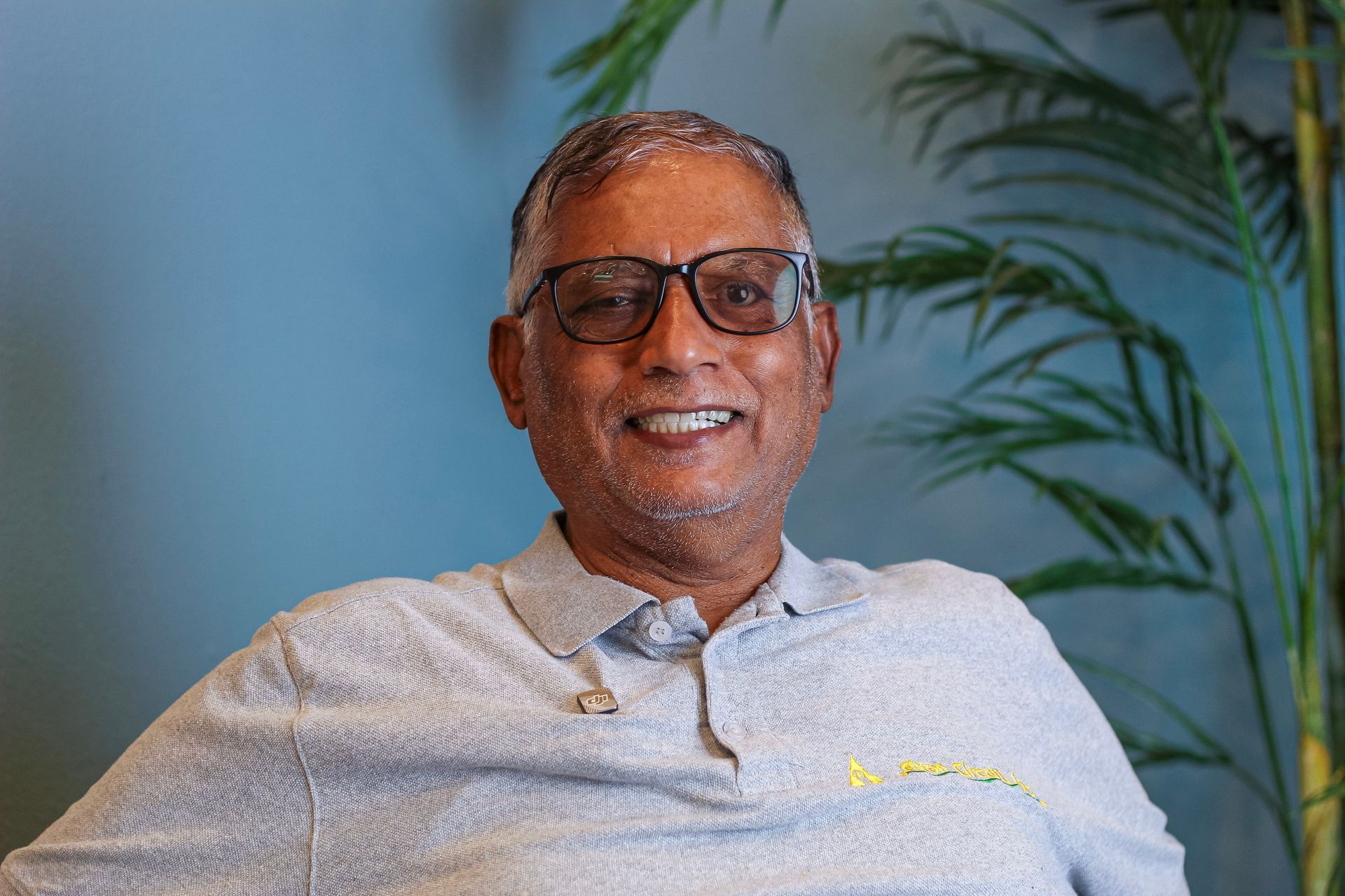 Smiling person in glasses and gray polo seated indoors against a blue wall with a plant in the background