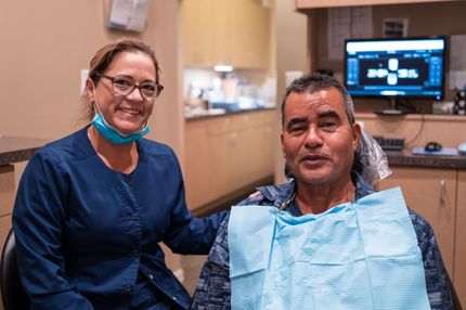Dentist in blue scrubs with patient in dental chair, both smiling at the camera. Dental office setting.