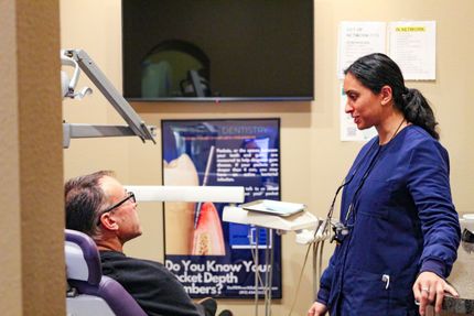 Dentist in blue scrubs consults with patient in dental chair; office setting.