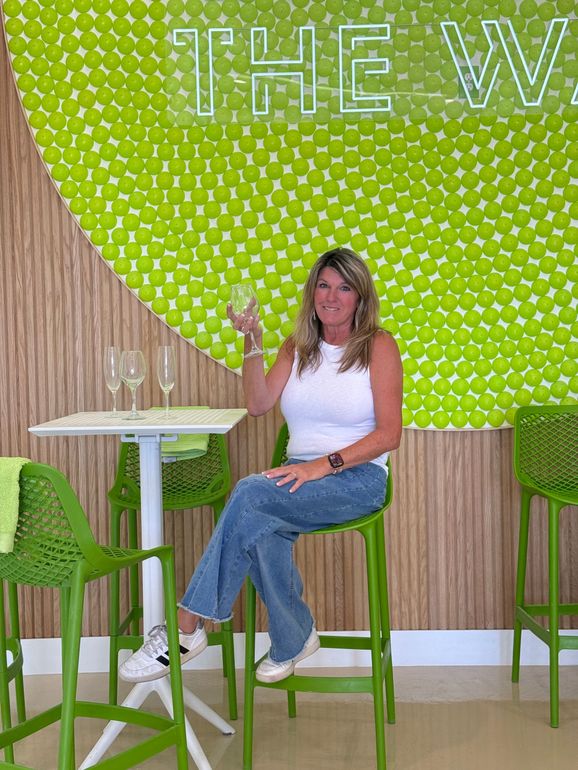 Woman seated at a high table in a green café, holding a drink against a leafy wall backdrop
