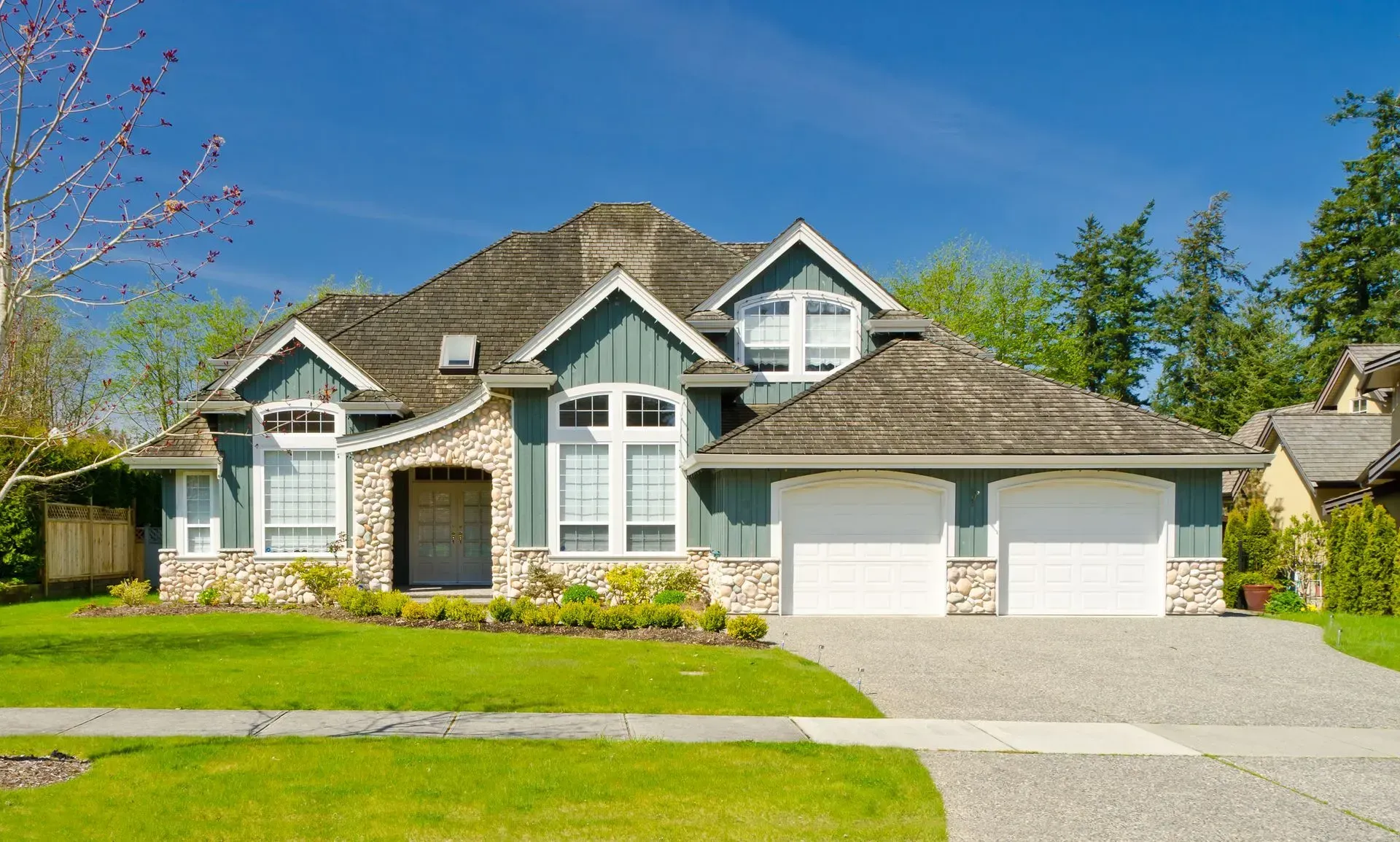 Suburban house with gray stone facade, white garage doors, and a green lawn under a blue sky