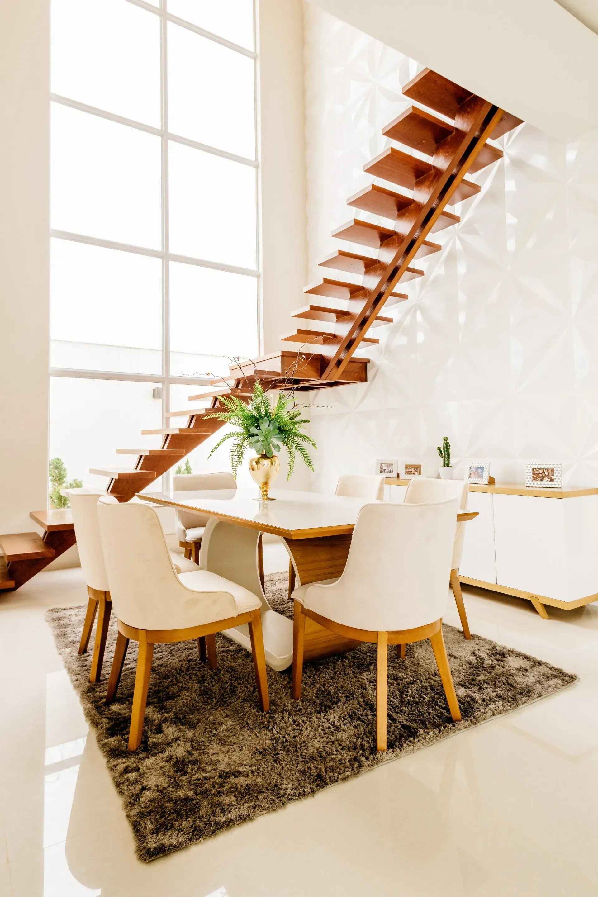 Bright modern dining area with wooden staircase, round table, cream chairs, and large windows