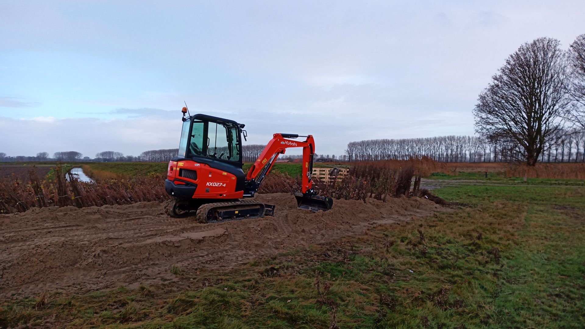 Een oranje graafmachine werkt op een modderig veld onder een bewolkte hemel.
