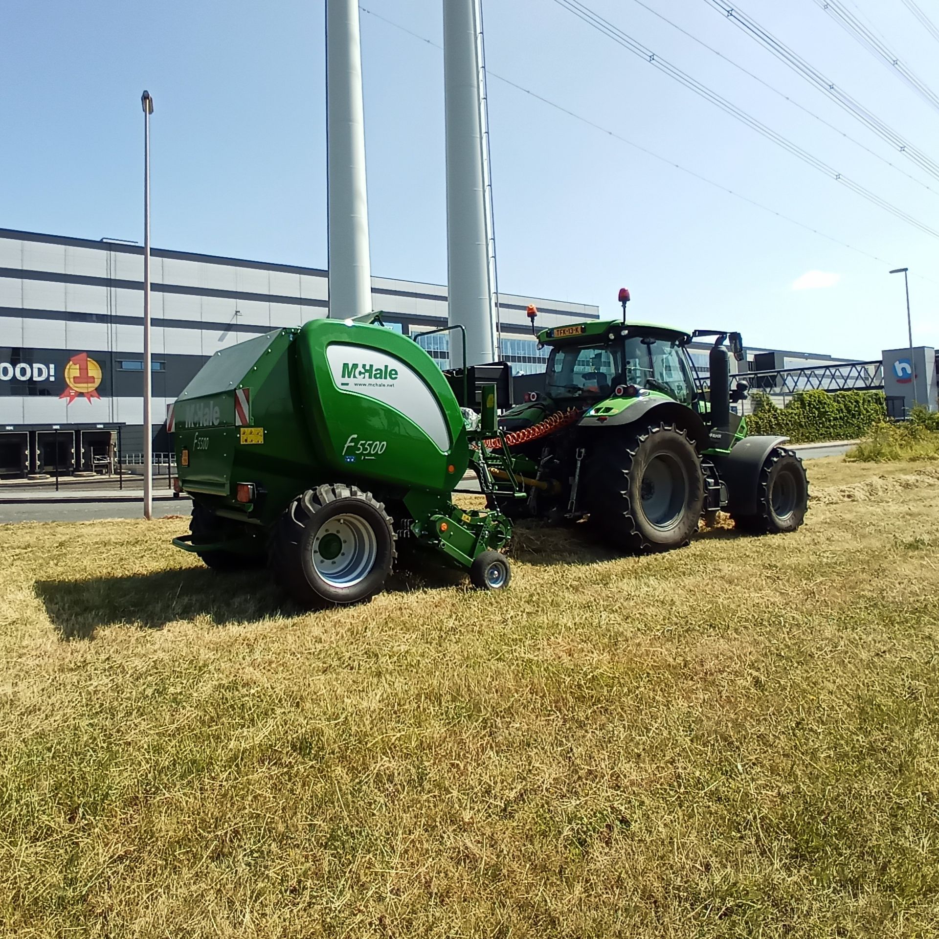 Groene tractor met aangekoppelde ronde balenpers geparkeerd op een grasveld nabij een weg en commerciële gebouwen.