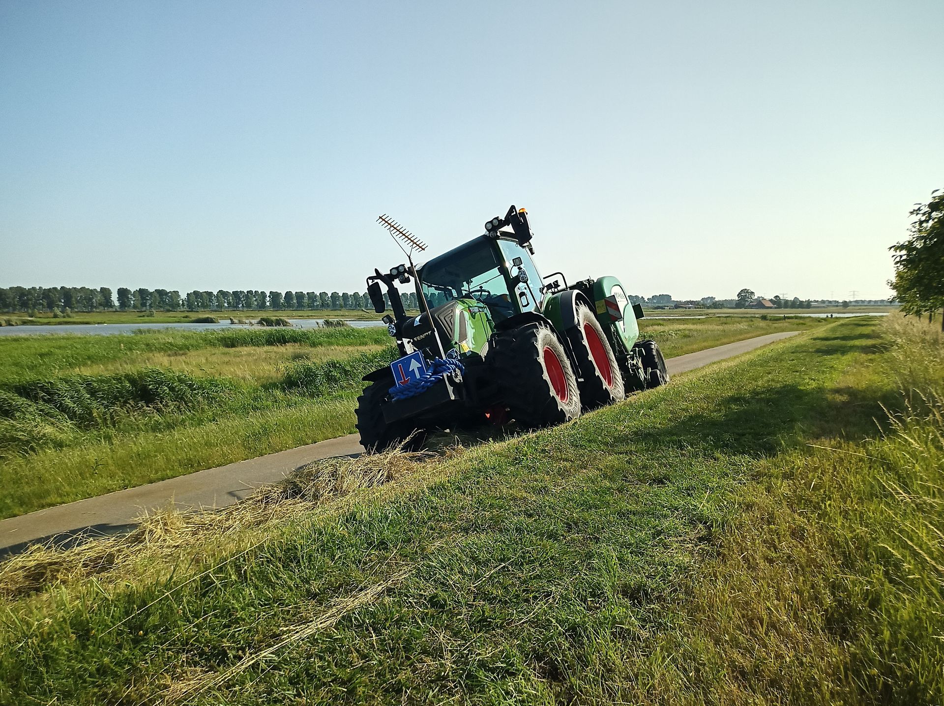 Een tractor staat scheef op een smalle landweg naast groene velden onder een heldere hemel.