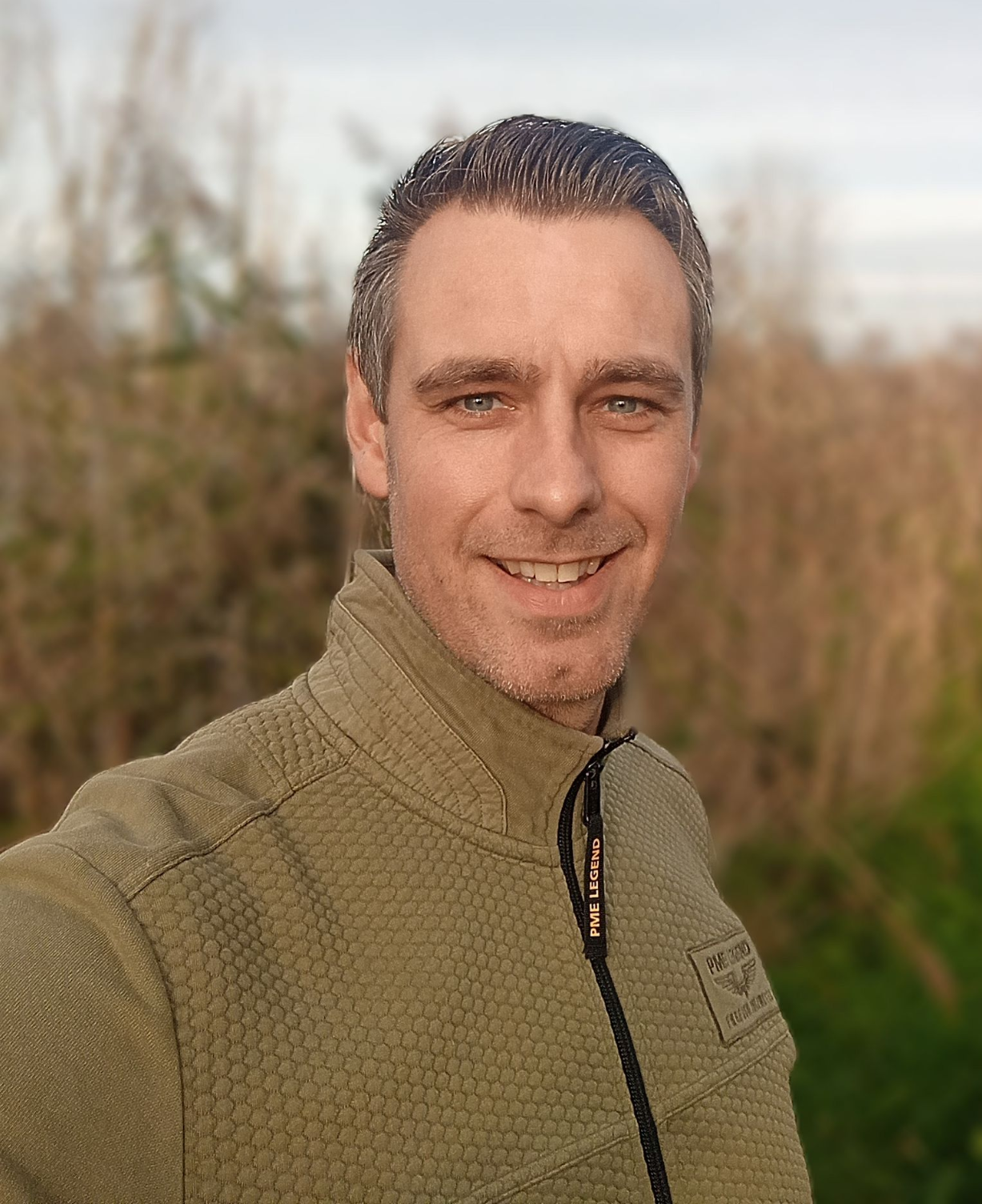 Een man lacht buiten in een beige jas, met wazige bomen op de achtergrond.