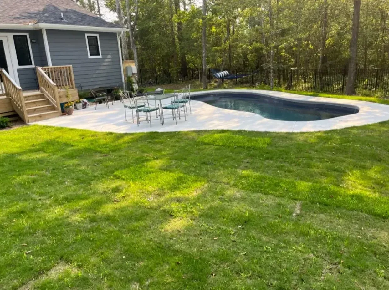 A grey house with a wooden deck overlooks a concrete patio, table set, and kidney-shaped pool in a green, grassy yard.