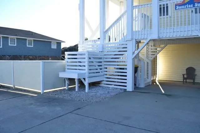 The base of a yellow stilt house featuring white slatted privacy screens, a gravel area, and a grey chair on a driveway.